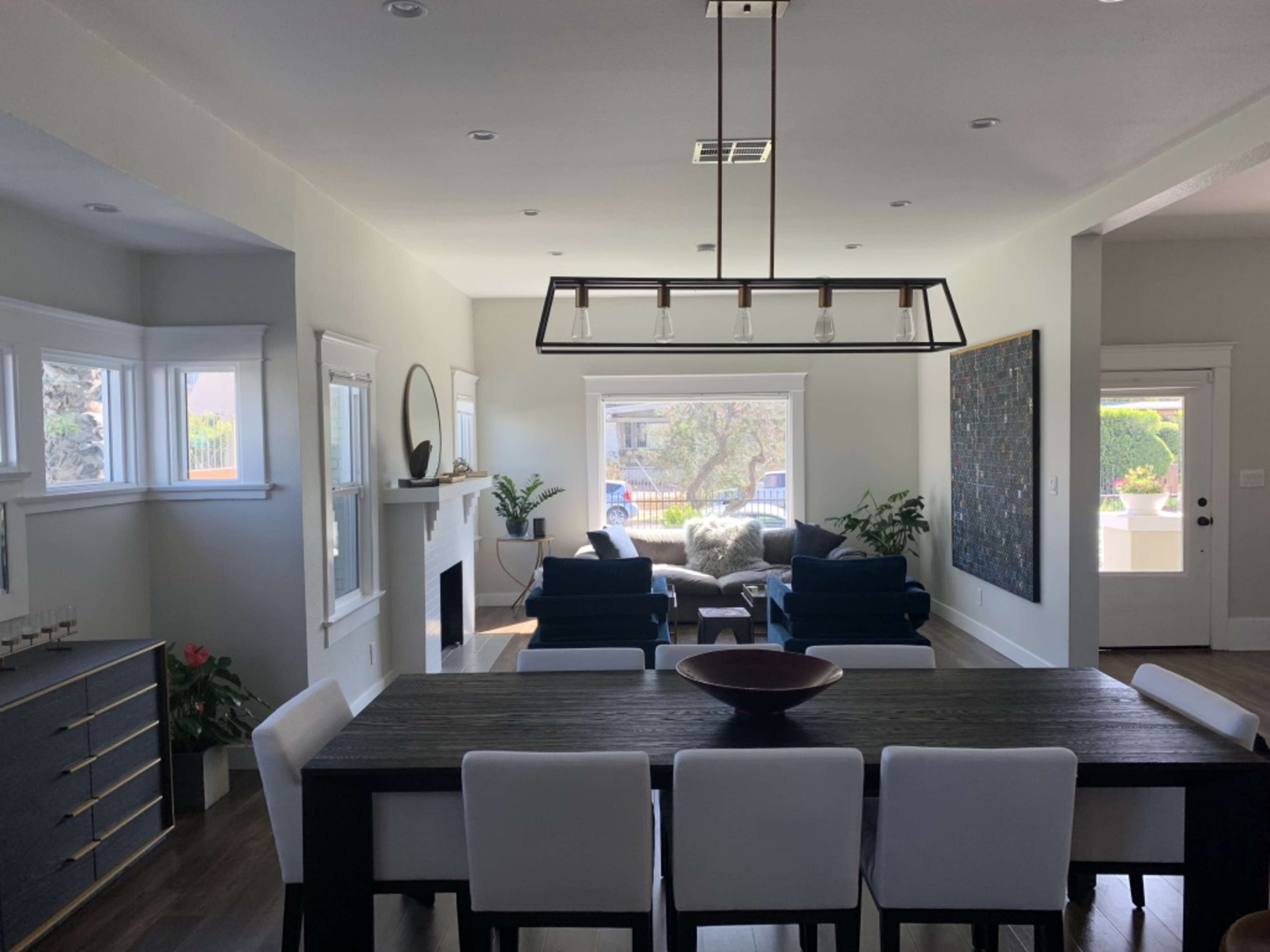 The image shows a modern living room and dining area with a dining table, chairs, a fireplace, and seating arranged near large windows.