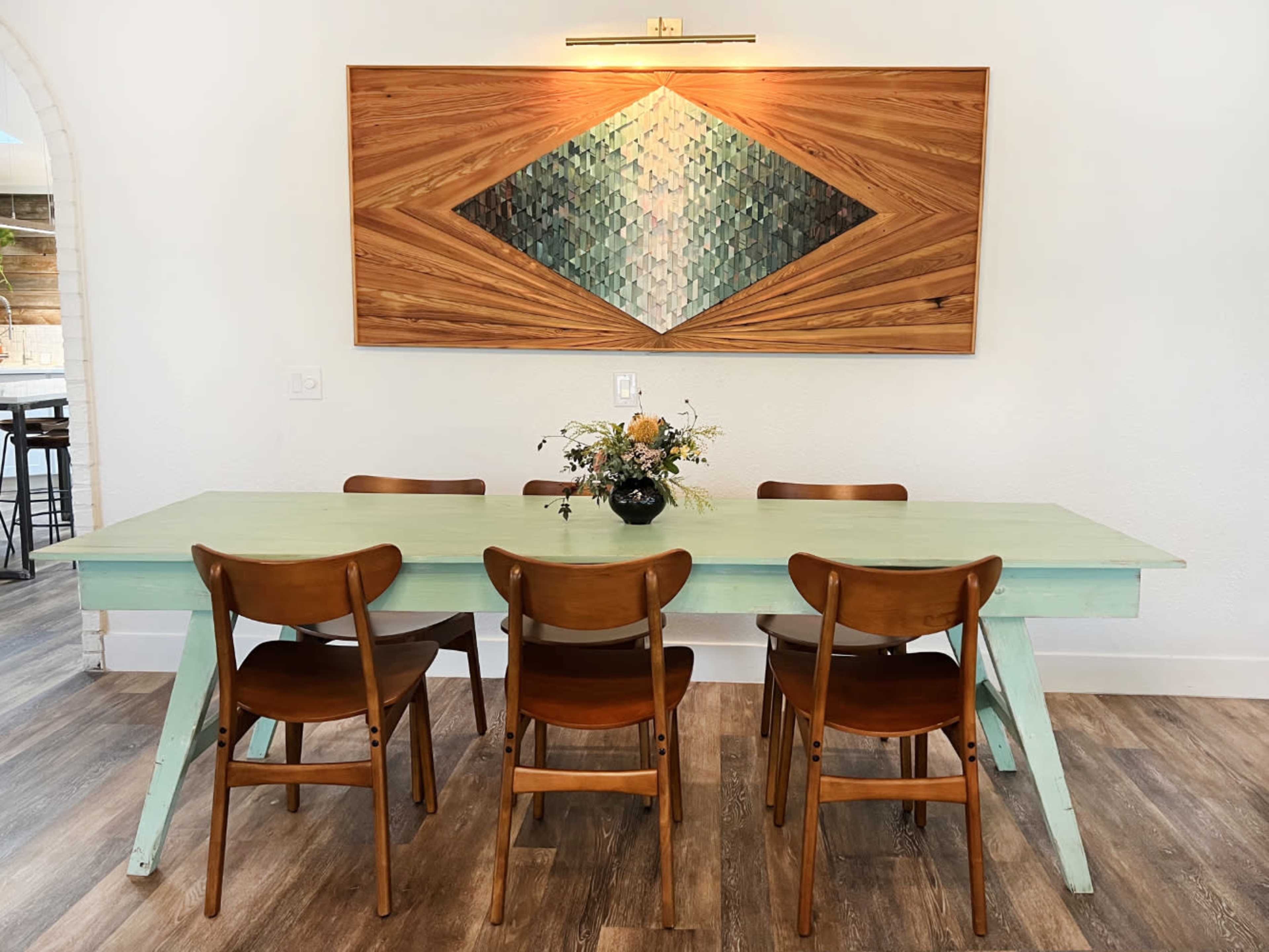 A light green table with six wooden chairs is set against a wall featuring a geometric wooden artwork.