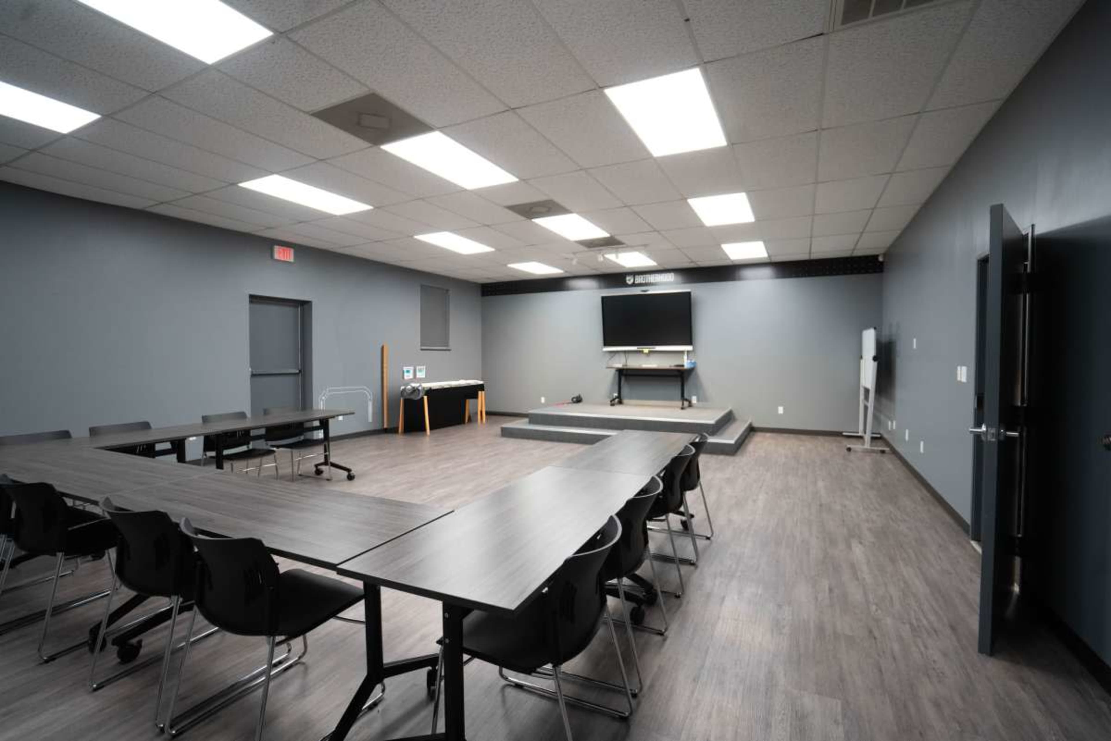 A spacious, empty classroom features multiple long tables and chairs arranged neatly, with a stage and a wall-mounted screen at the front.
