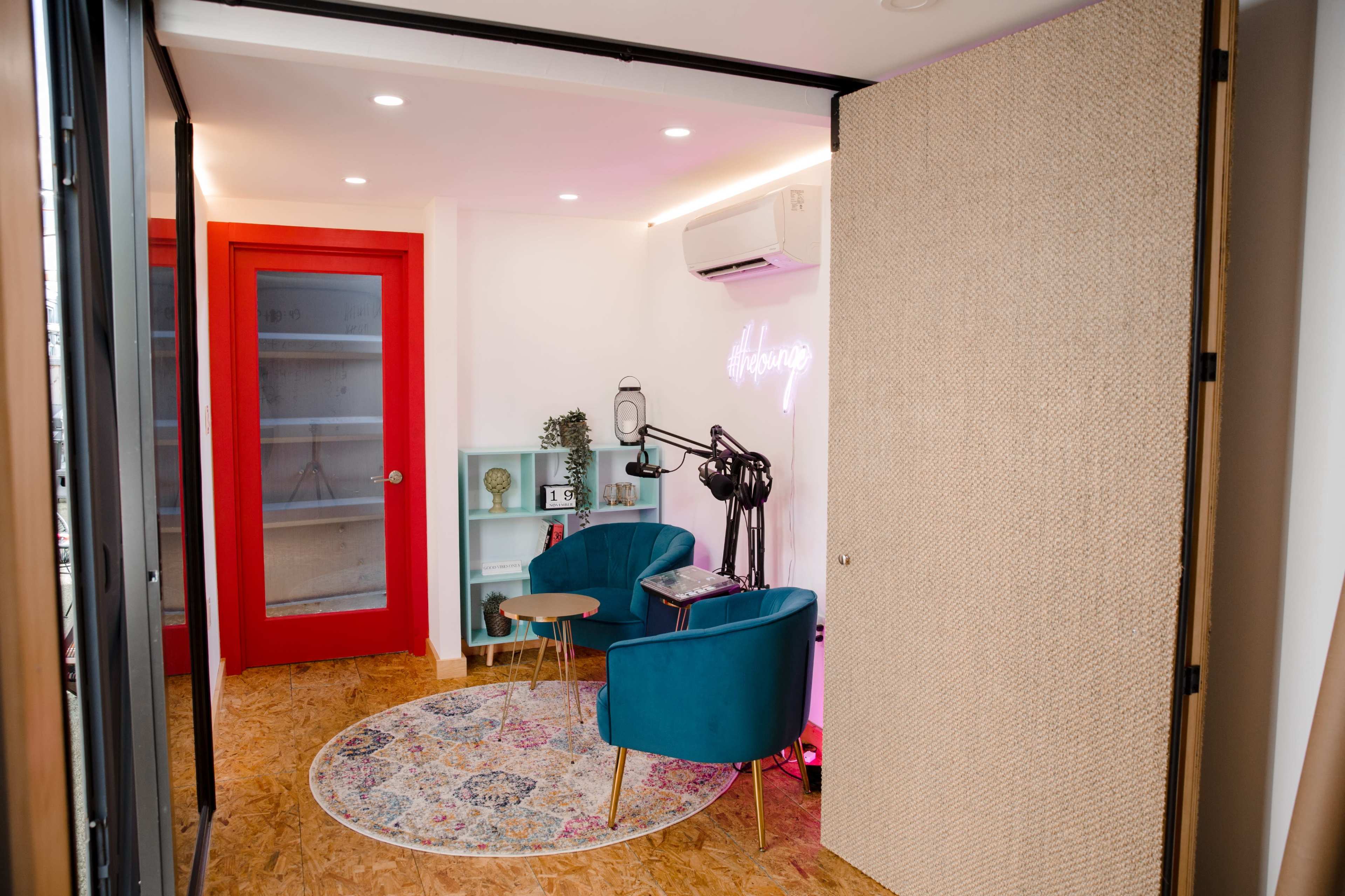 The image shows a modern room with two teal chairs, a small wooden table, and a colorful rug, featuring a red glass door and decorative shelves.