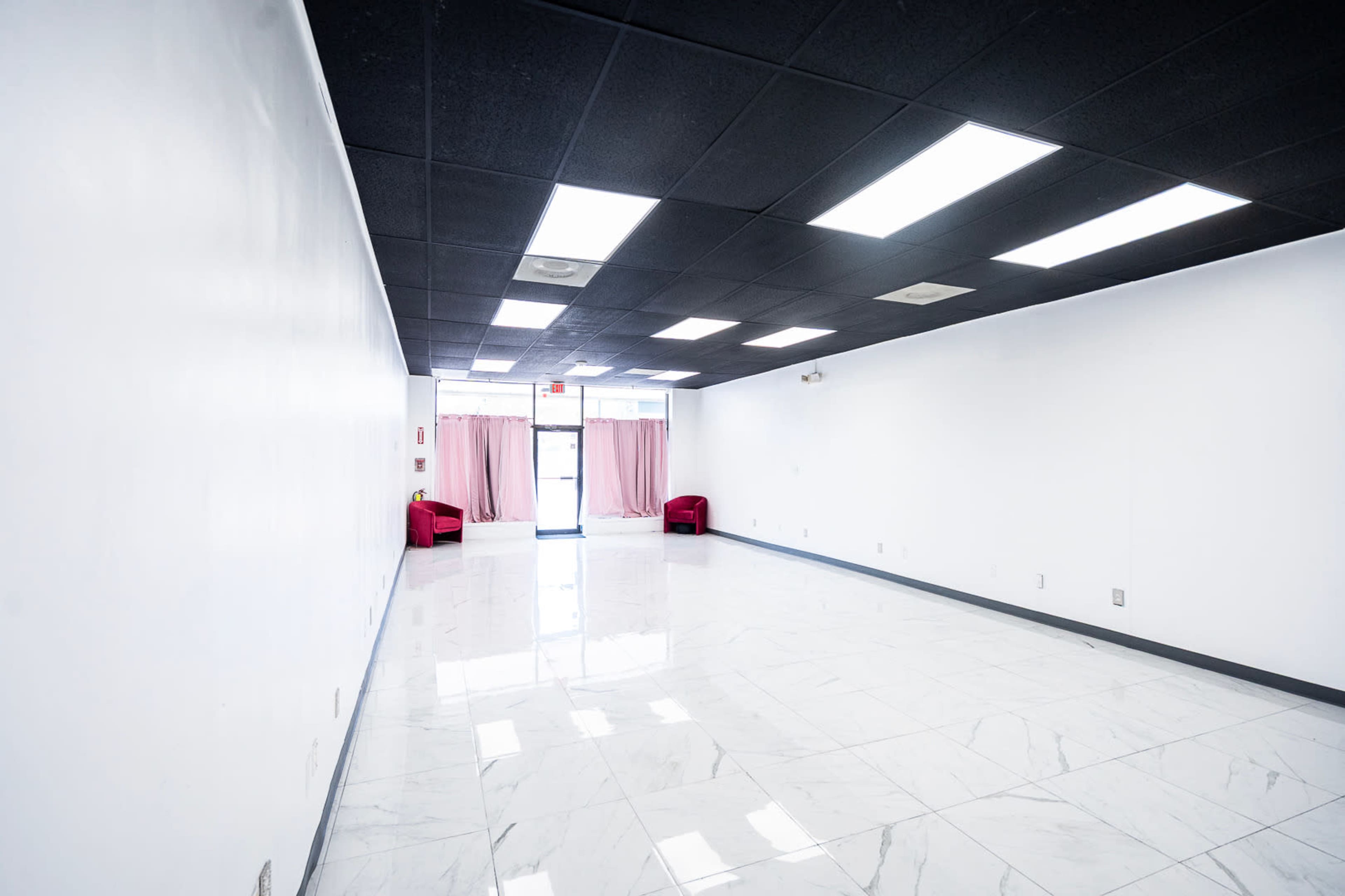 The image shows a vacant room with white walls, glossy tile flooring, and two red chairs positioned near a doorway with pink curtains.