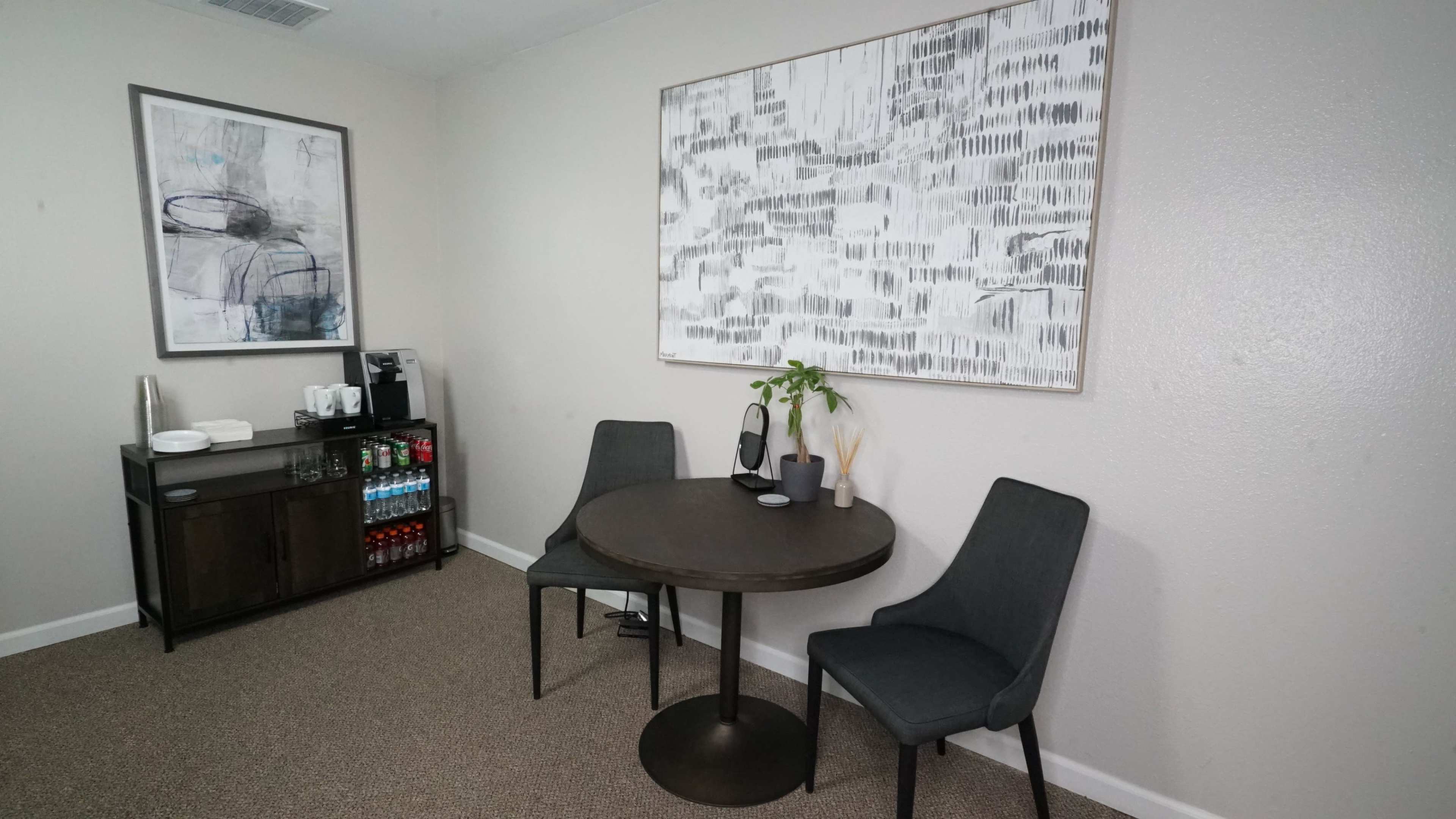 A small dining area features a round table with two gray chairs next to a sideboard stocked with beverages and a large abstract painting on the wall.