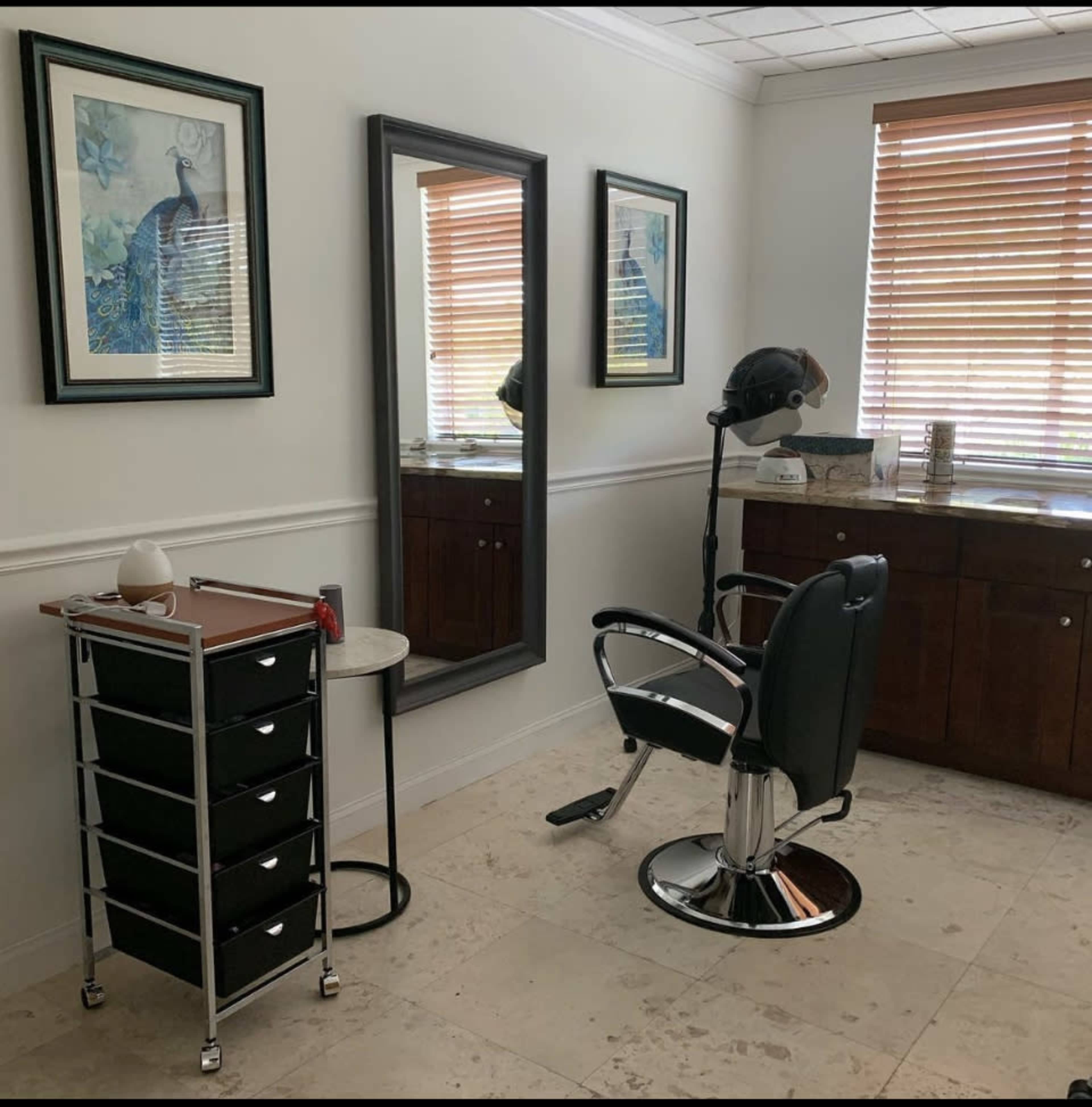 A hair salon room features a black salon chair, a mirror, a storage cart with drawers, and framed artwork on the walls.