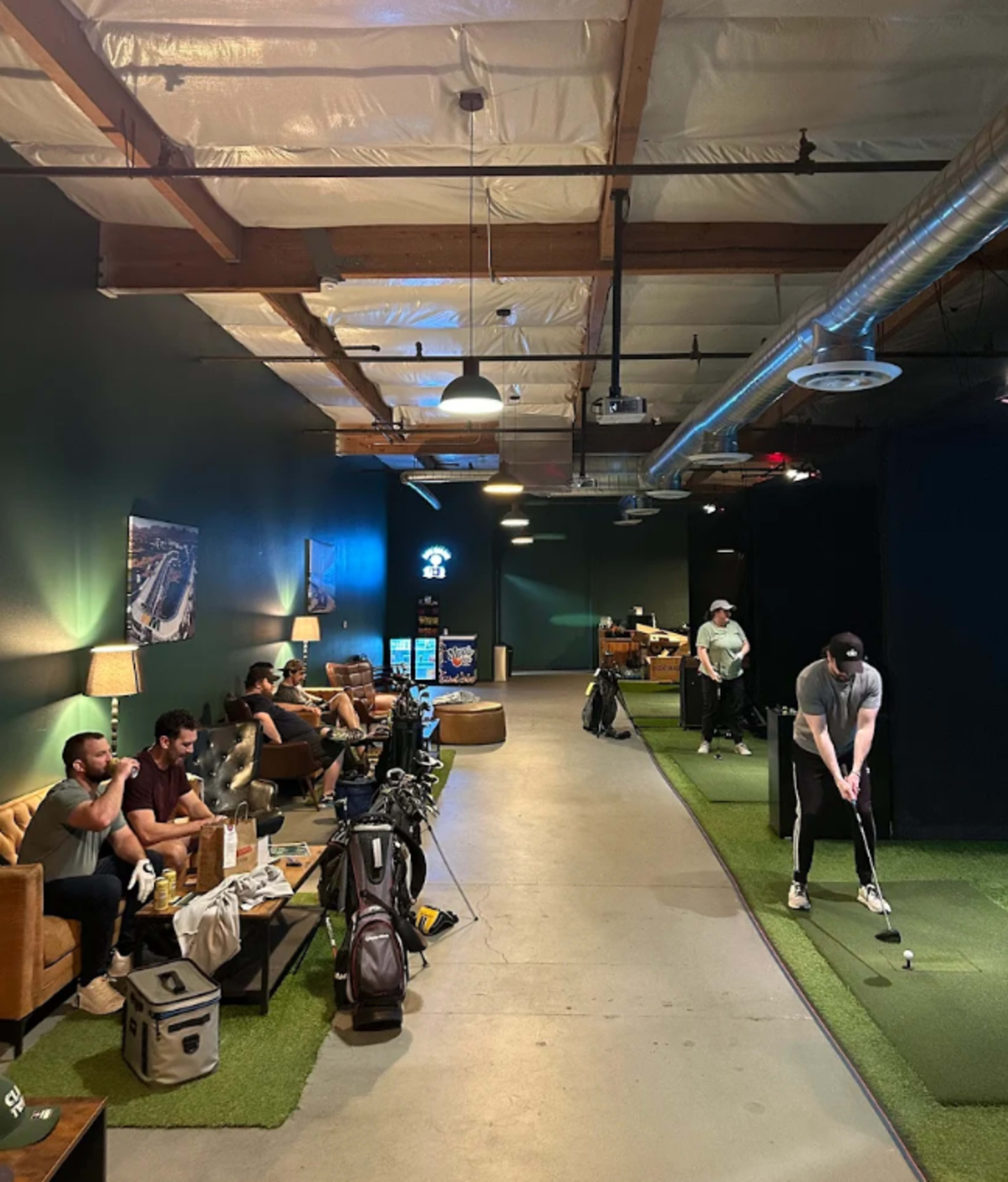 The image shows a dimly lit indoor golfing facility with several people engaged in various activities, including one person putting on a green mat while others are seated on sofas.