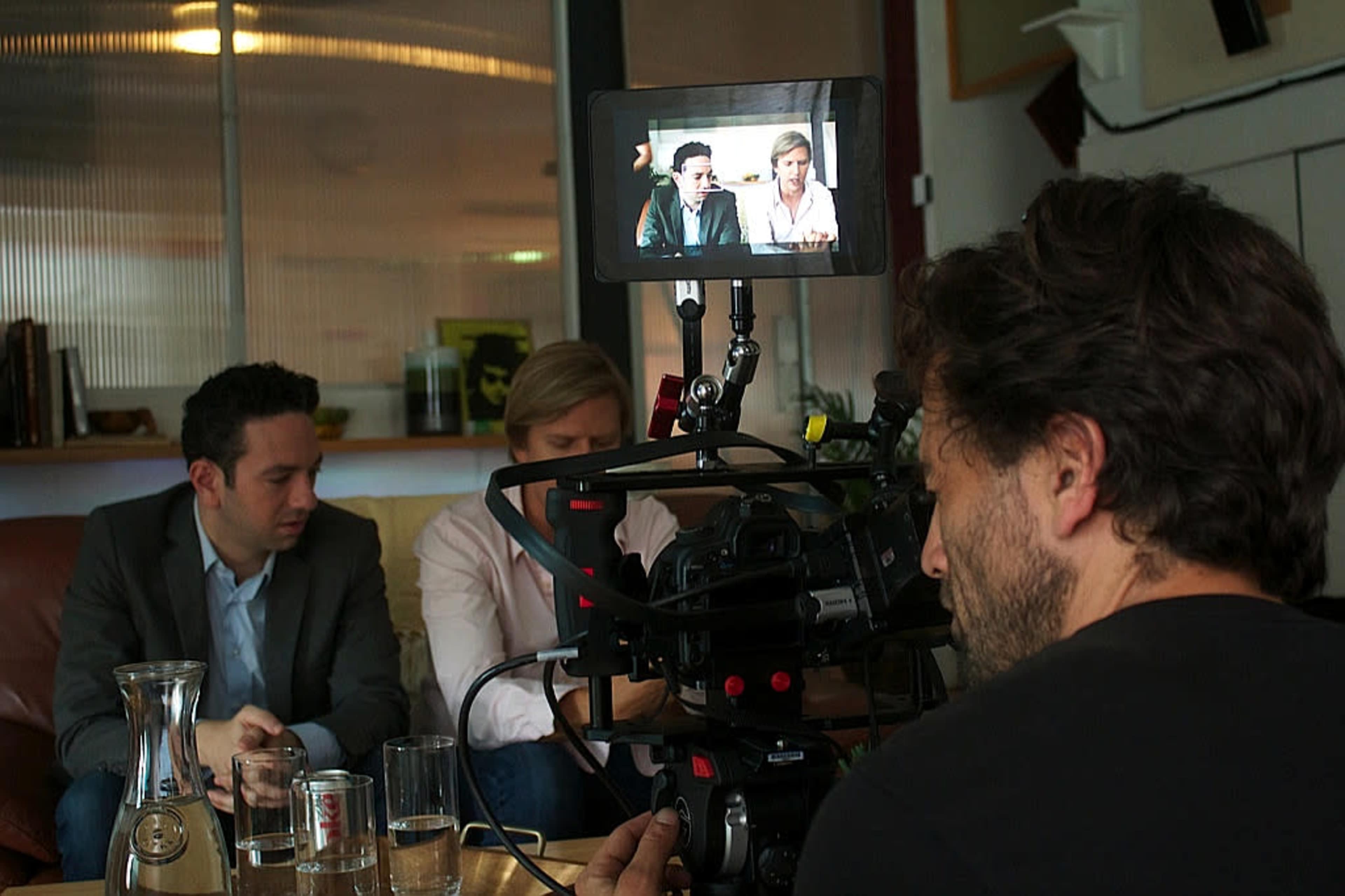 A camera operator films two men seated at a table during an interview in a well-lit indoor setting.