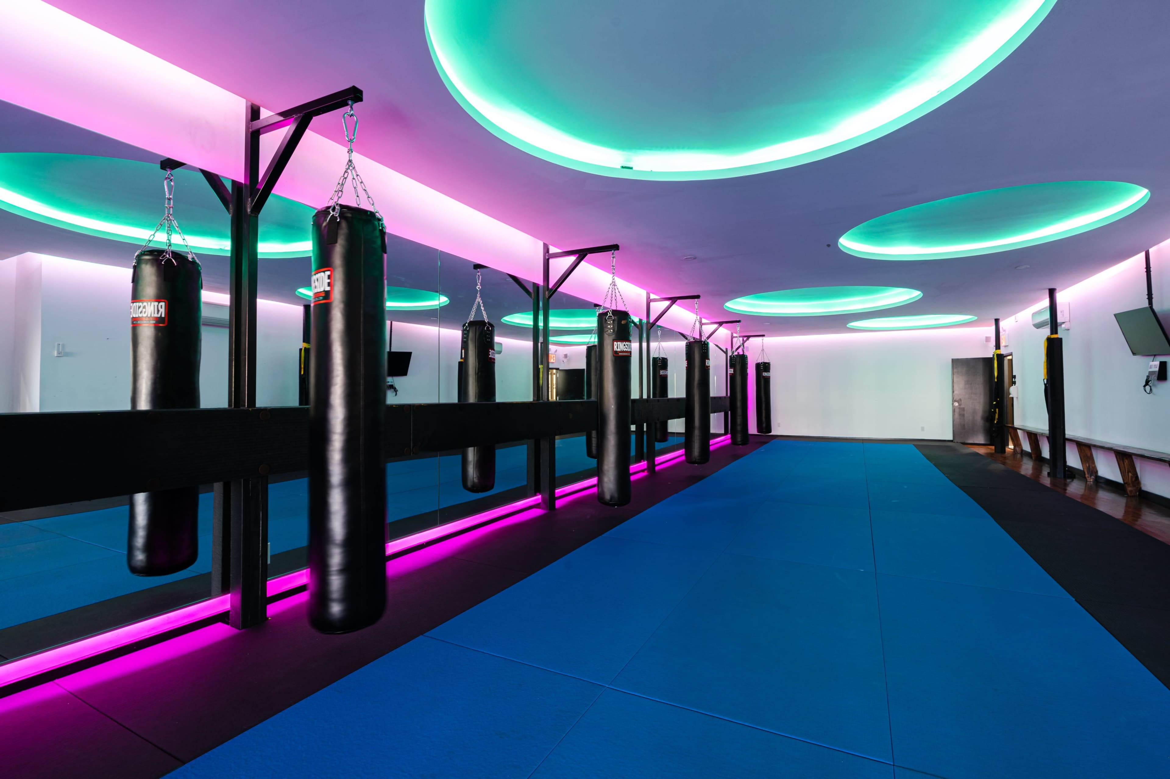 A modern boxing gym with black punching bags hanging from a black frame under circular, colorful ceiling lights and a blue mat on the floor.