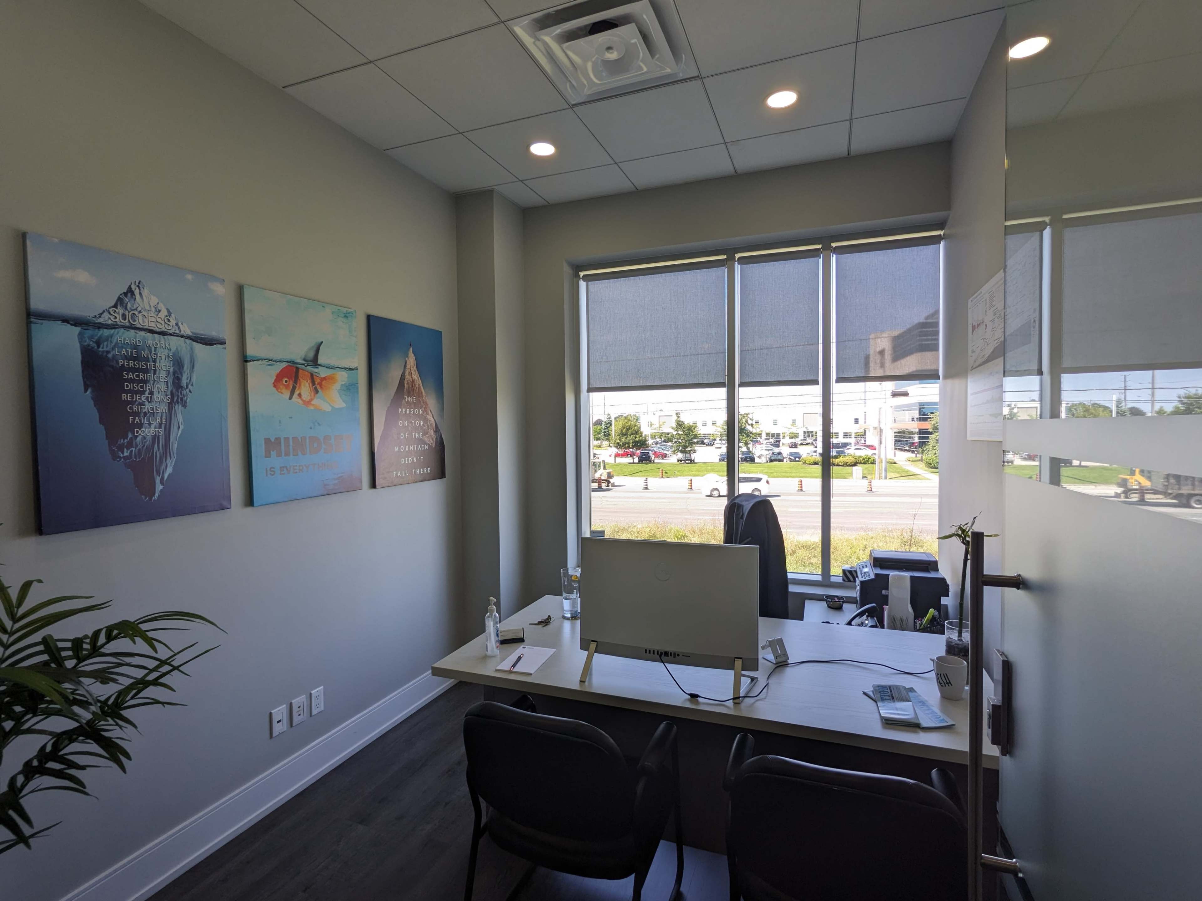 A well-lit office space with a desk, computer, and three framed motivational posters on the wall, near a large window.