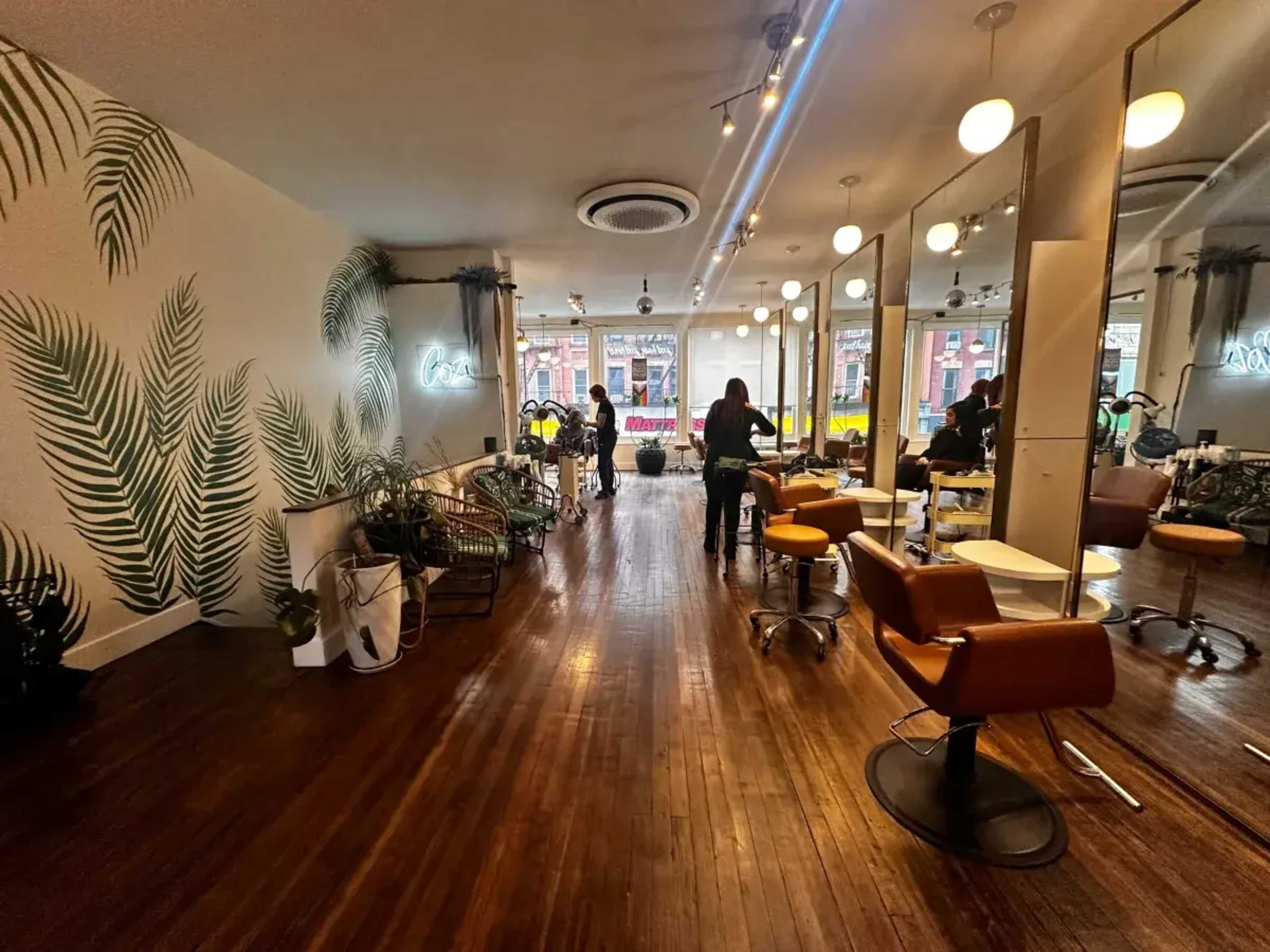A hair salon features wooden flooring, large mirrors, and plants, with stylists attending to clients.