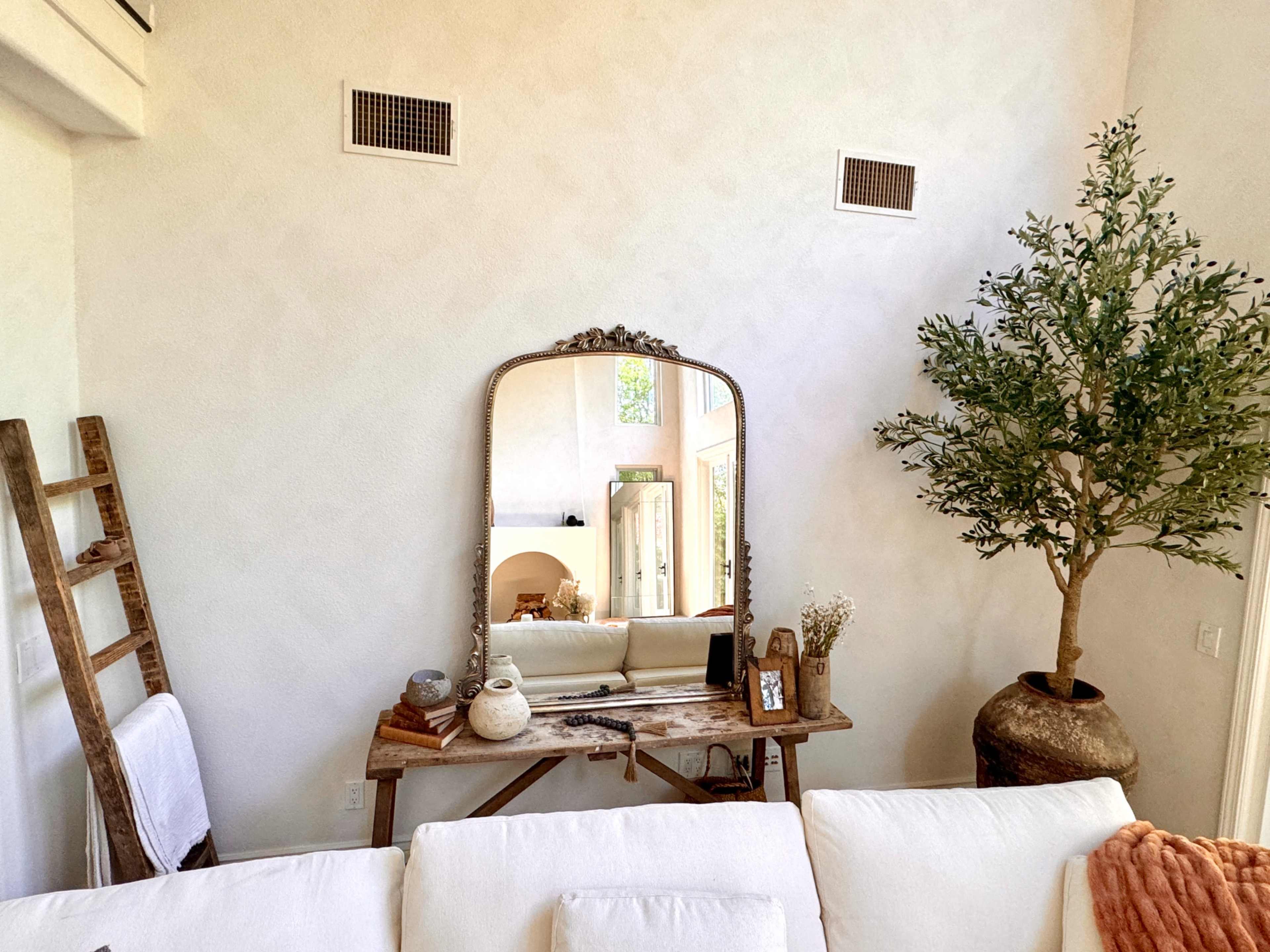 A mirror is positioned above a rustic table adorned with decorative items and a plant, beside a white couch and a wooden ladder against a textured wall.