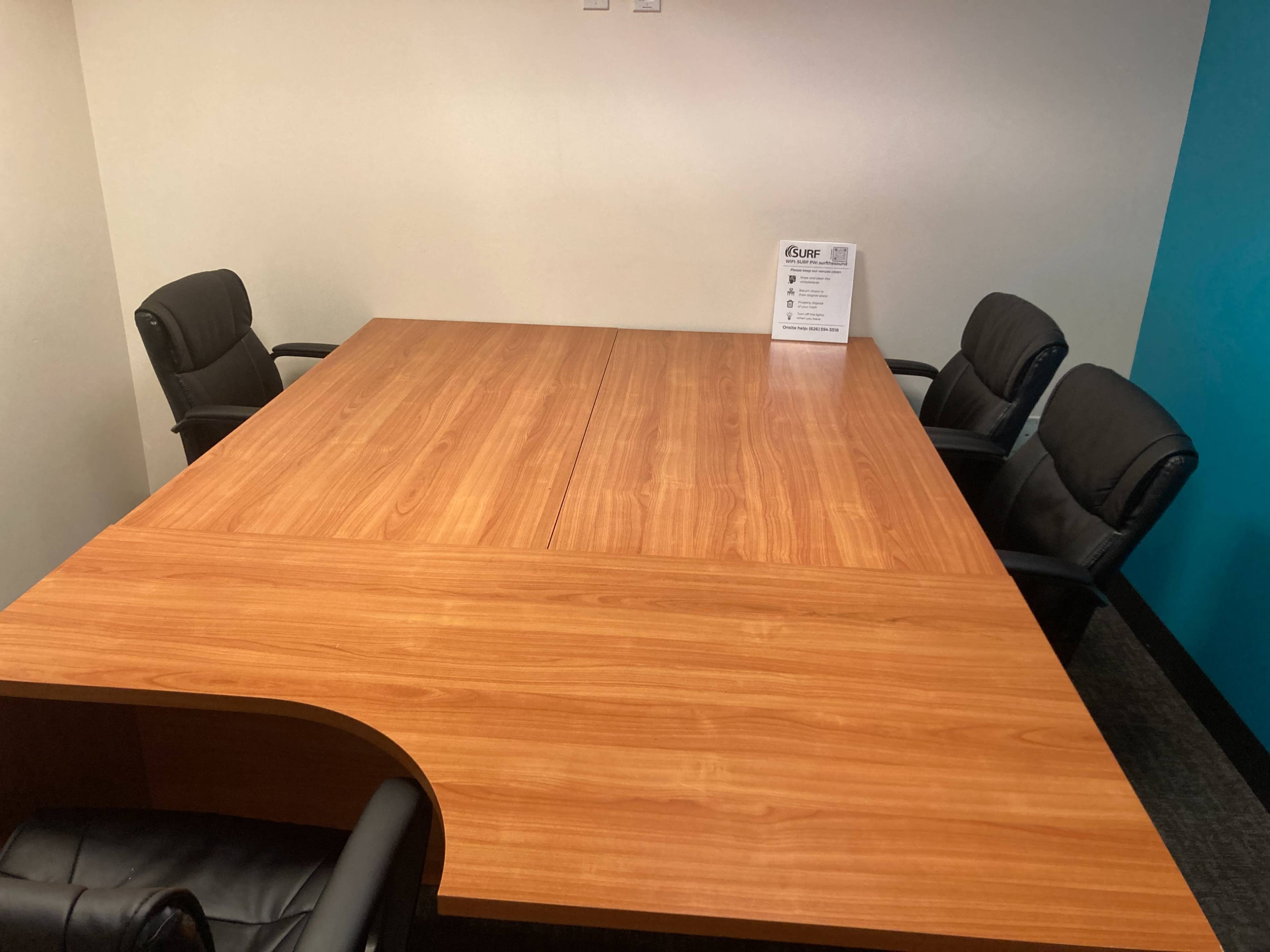 A wooden conference table sits in a small meeting room with four black chairs arranged around it.