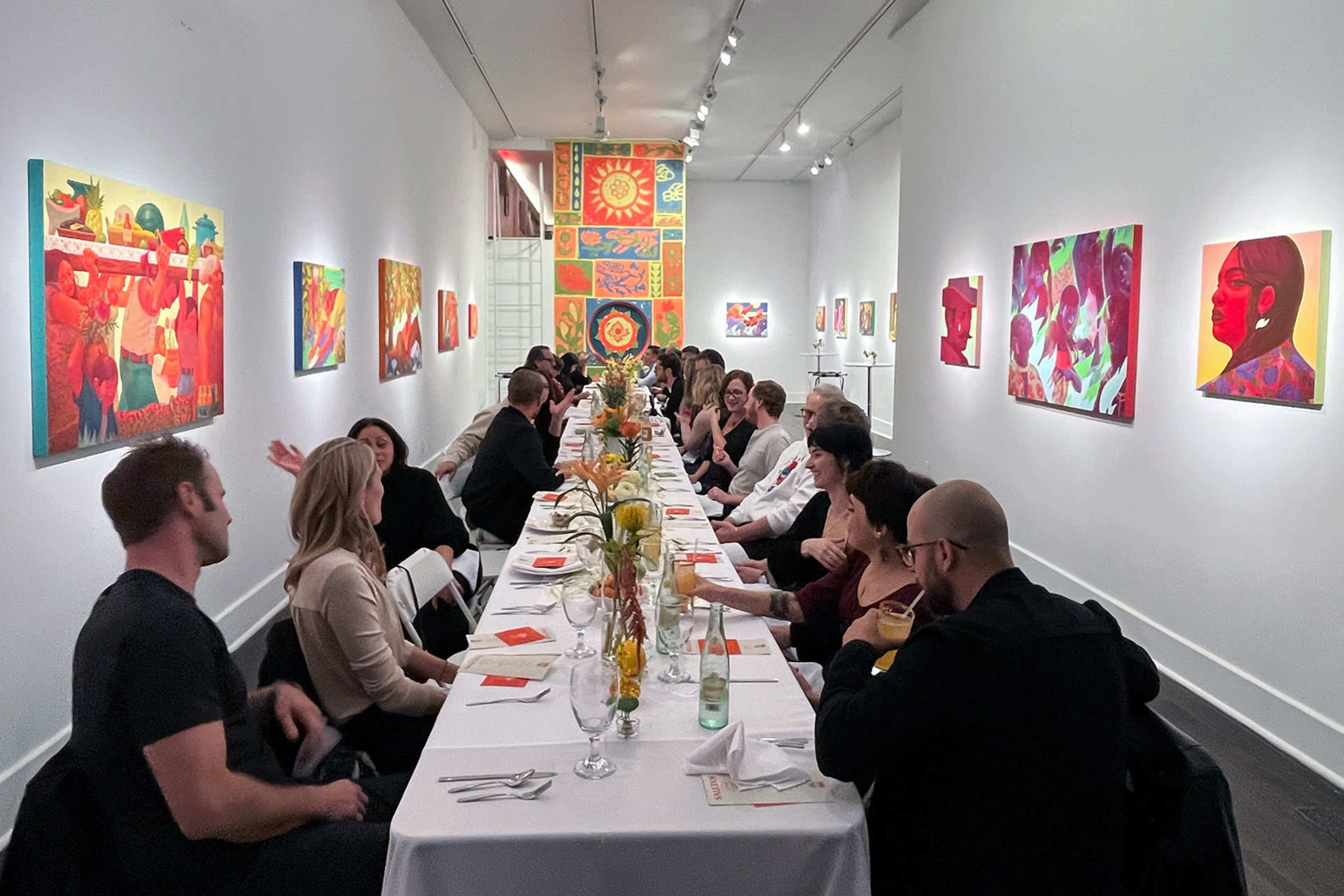 A long dining table is set with white tablecloths in an art gallery, surrounded by colorful artwork on the walls and filled with guests engaged in conversation.