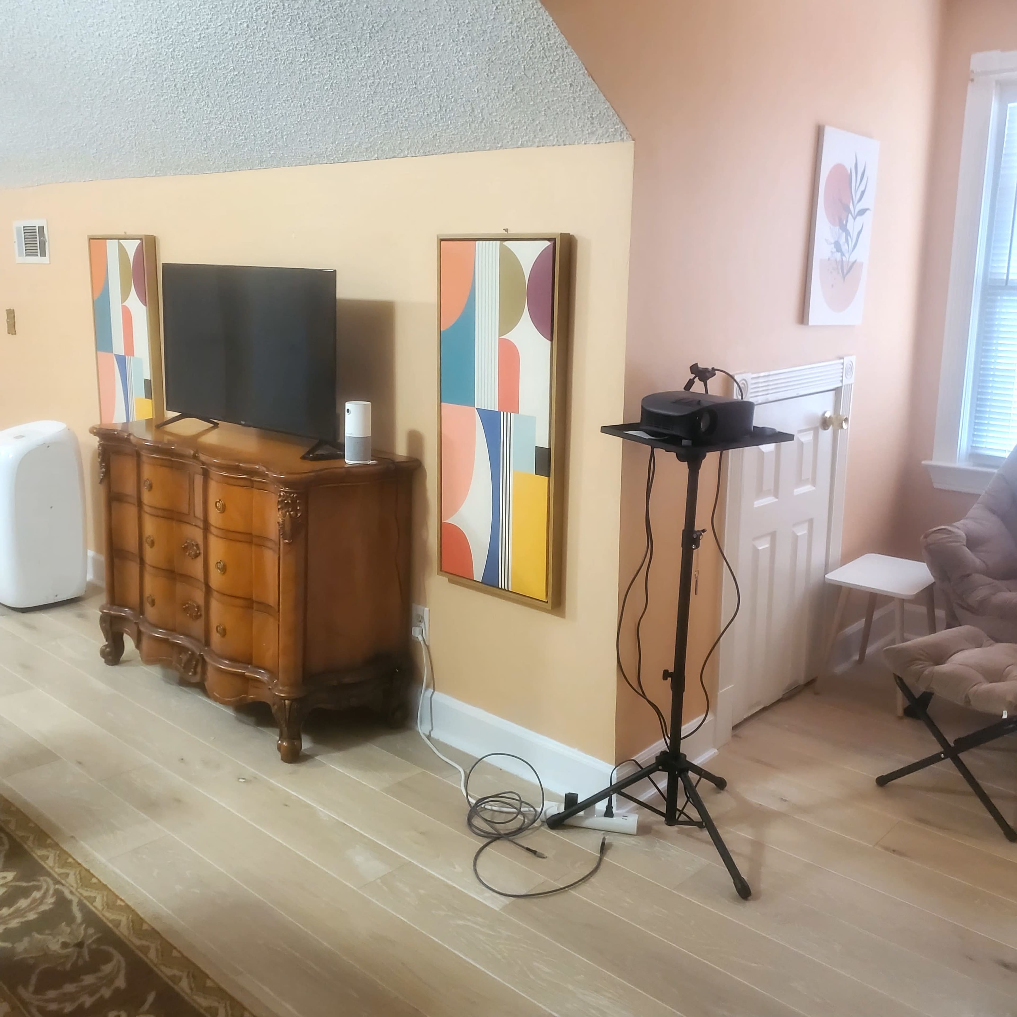 A room with a wooden TV stand, a television, two colorful abstract paintings on the walls, and a folding chair next to a small table with a device on a stand.