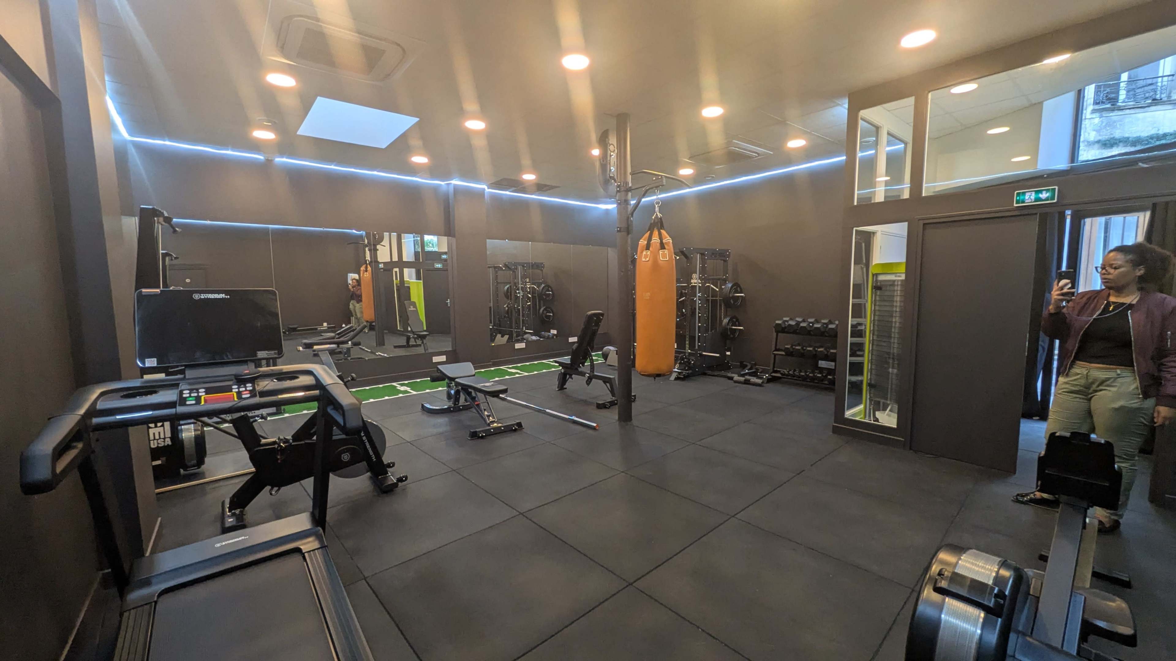 The image depicts a modern gym with various workout equipment, including a treadmill, weightlifting area, and a boxing bag, all arranged on rubber flooring.
