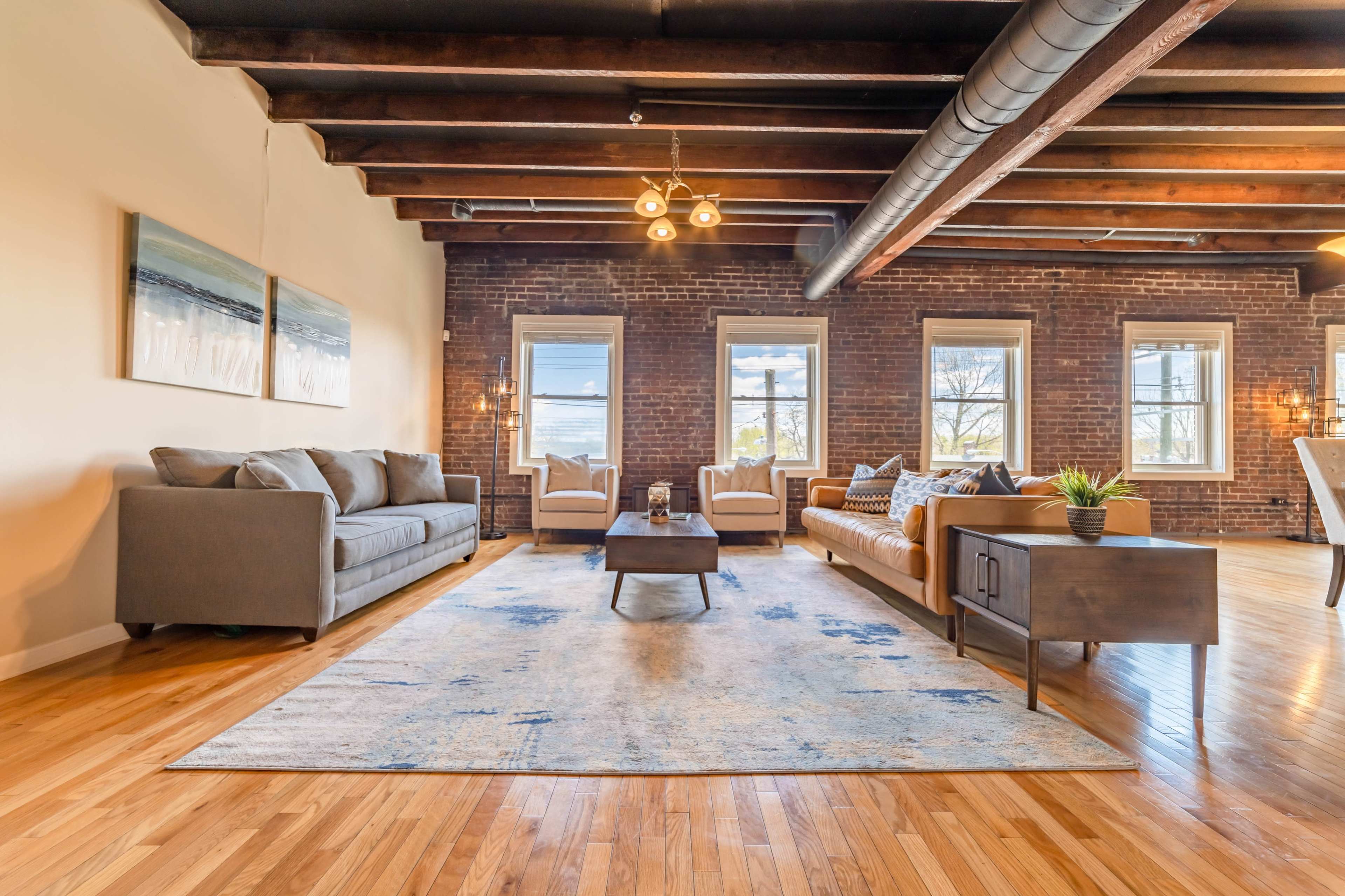 A modern living room features a spacious arrangement of a gray sofa, a patterned couch, a wooden coffee table, and a decorative rug under exposed beams and large windows.