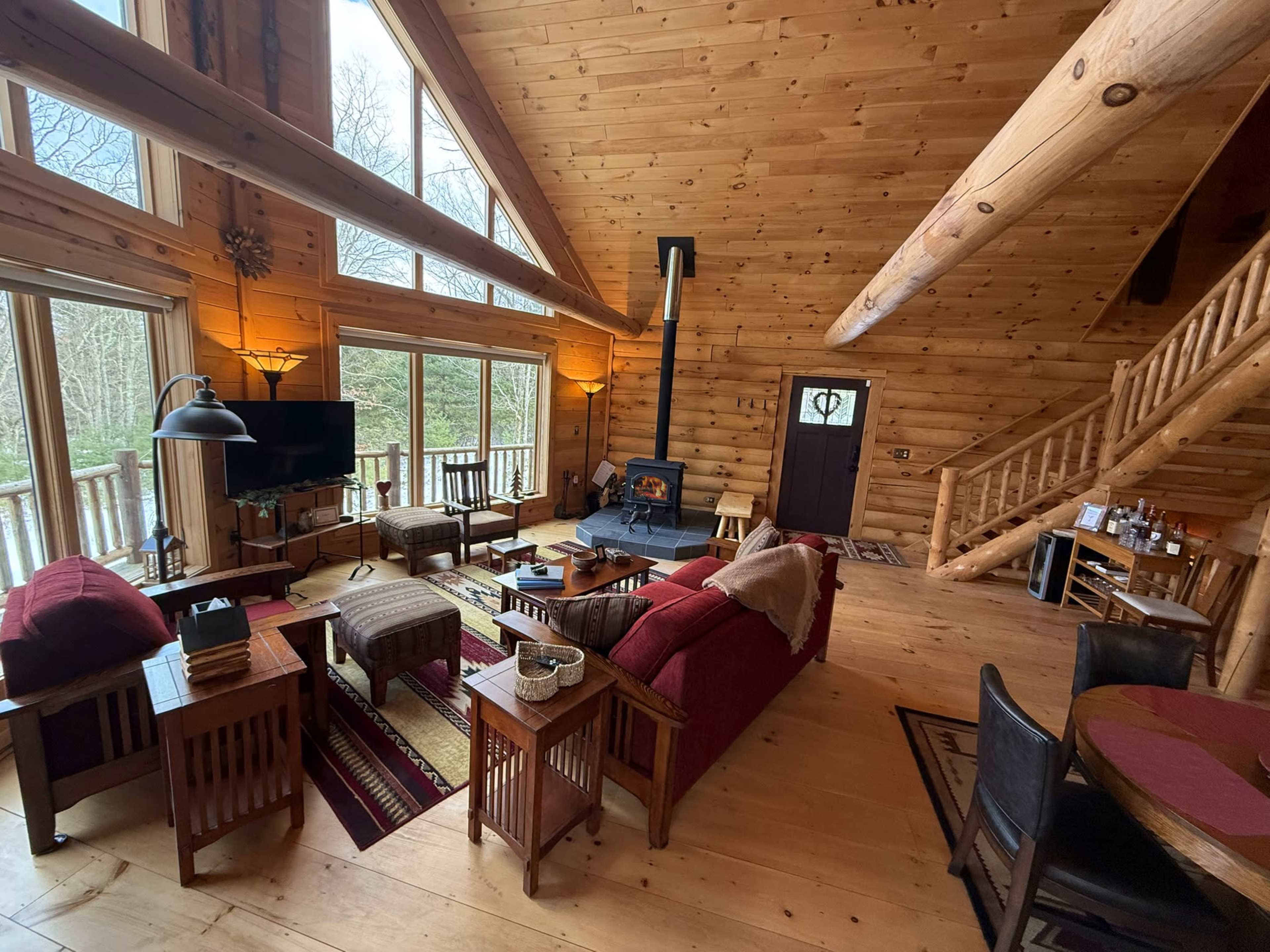 The interior of a rustic wooden cabin features a living area with a red sofa, wooden furniture, a stove, and large windows overlooking a forested view.