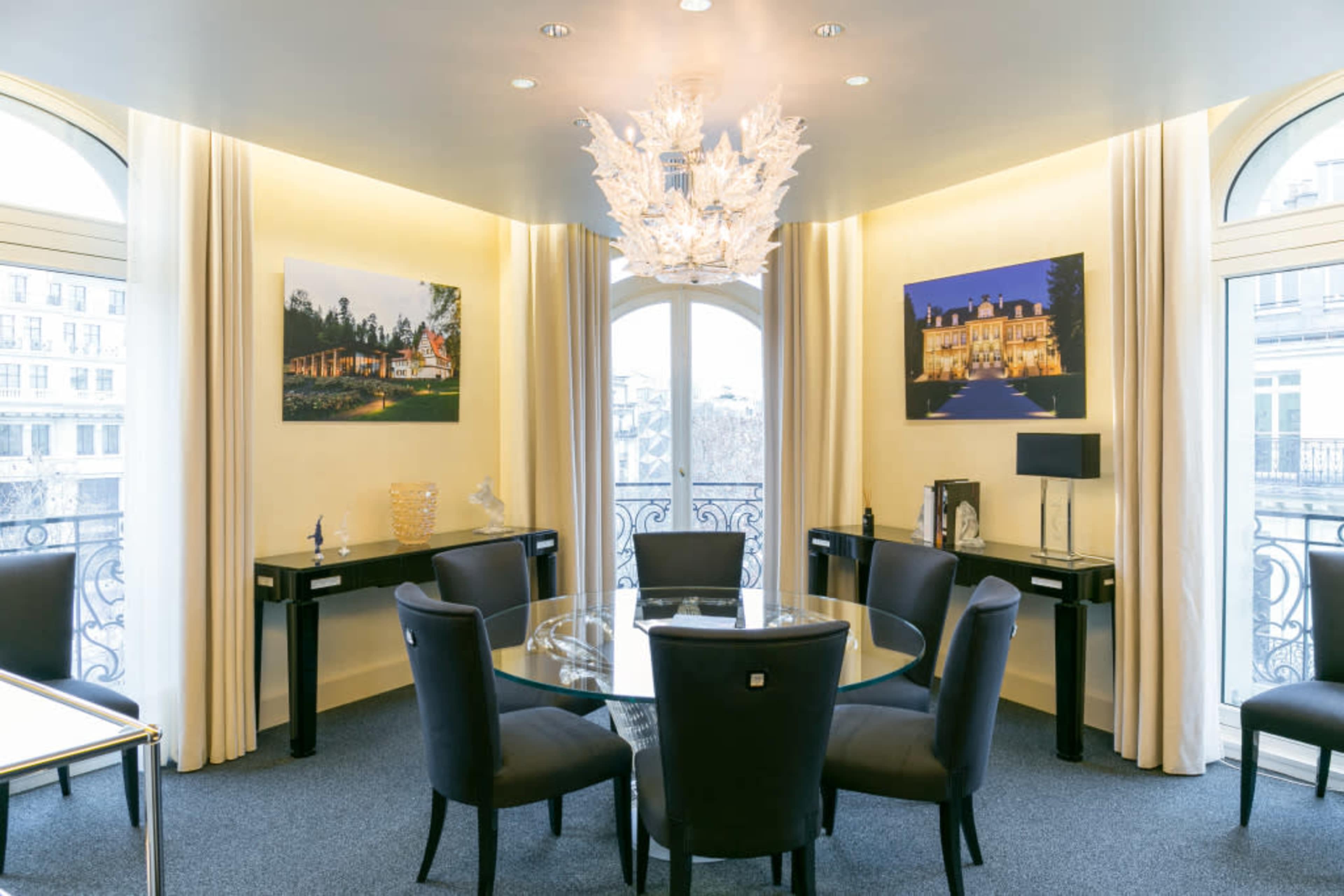 A modern meeting room with a glass table surrounded by black chairs, featuring large windows and two framed photographs on the walls.