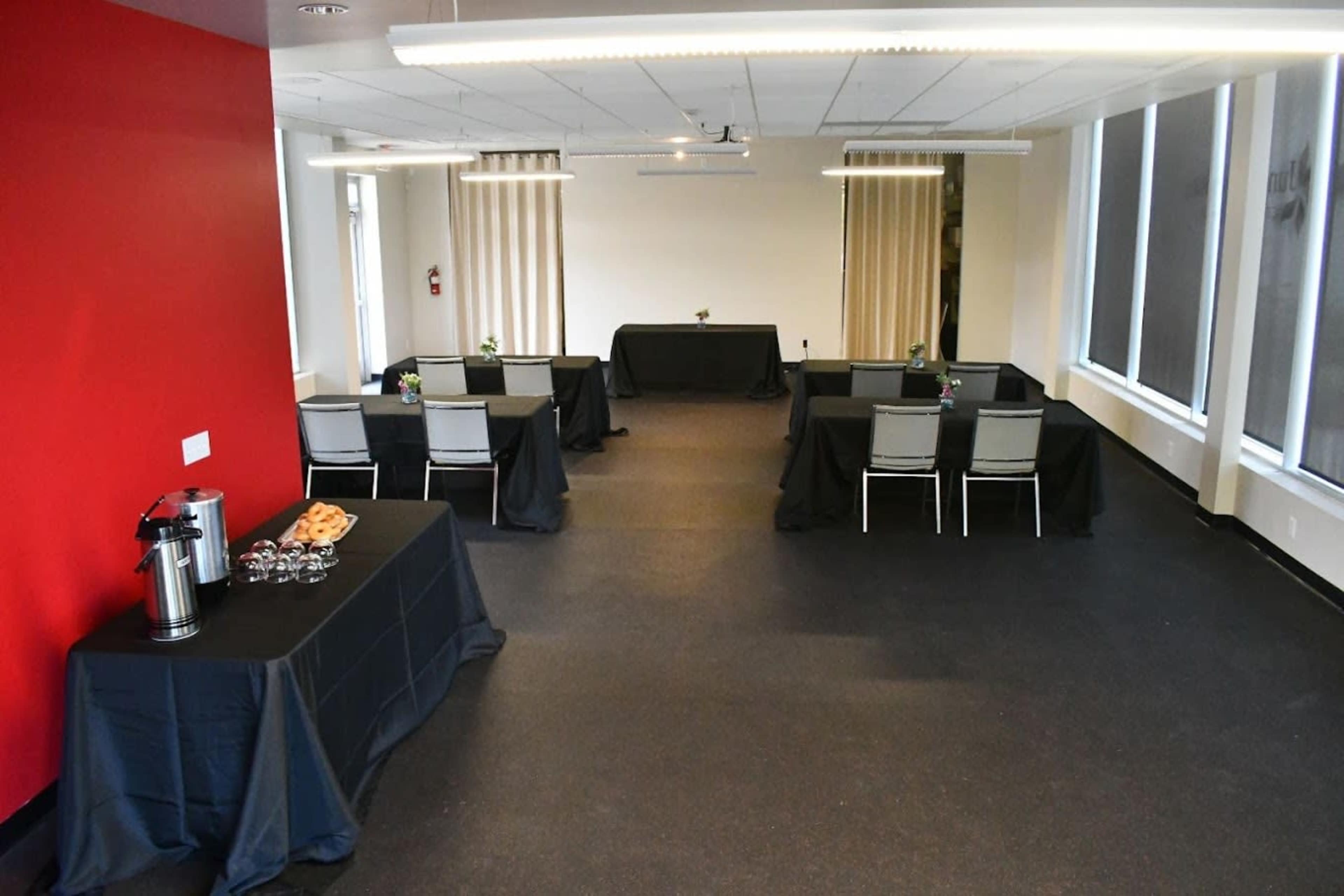 A conference room set up with several tables covered in black tablecloths, some with refreshments and floral centerpieces, against a backdrop of a red wall.