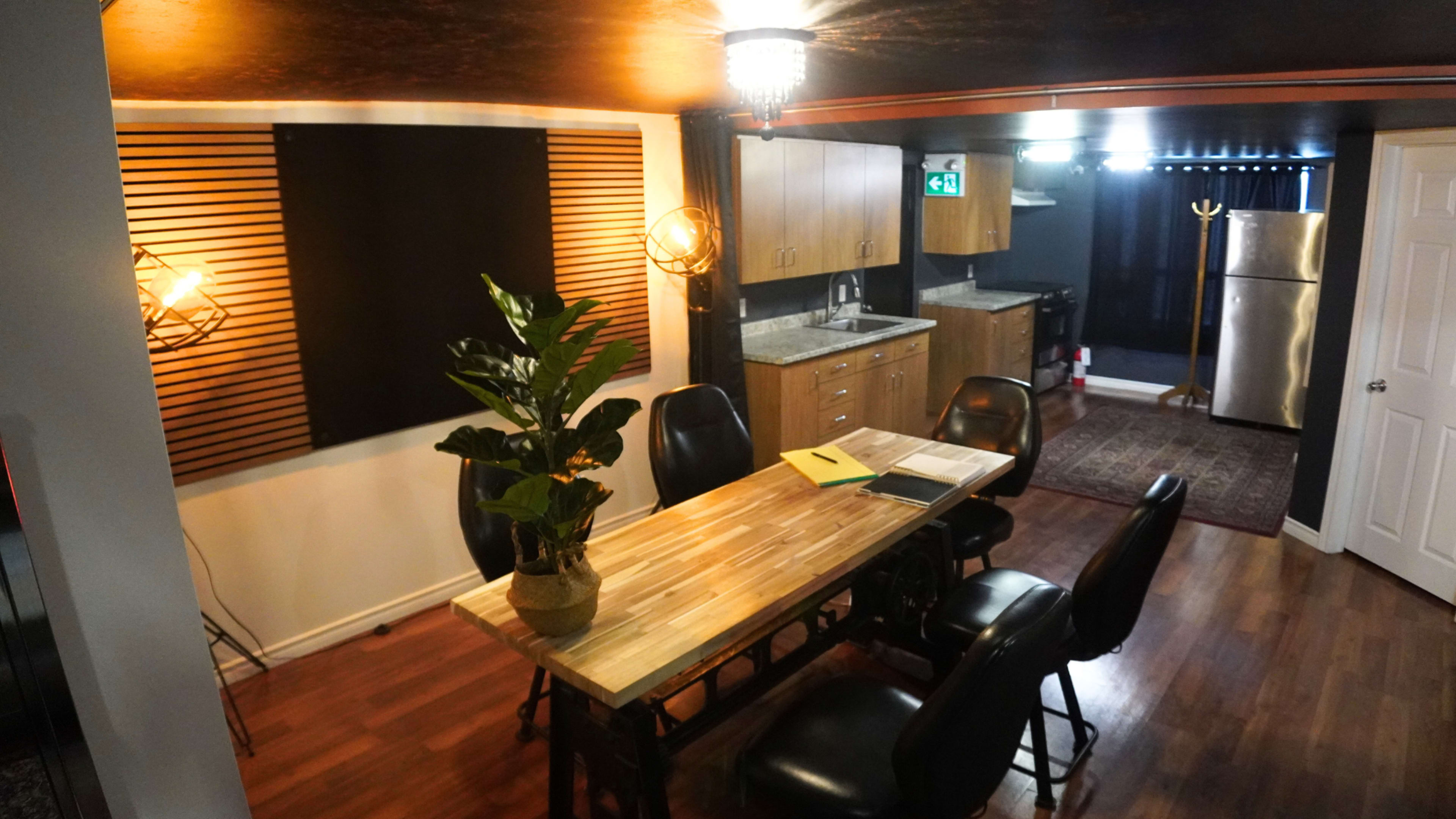 The image shows a modern kitchen and dining area featuring a wooden table with black chairs, a potted plant, and a kitchenette with appliances in the background.