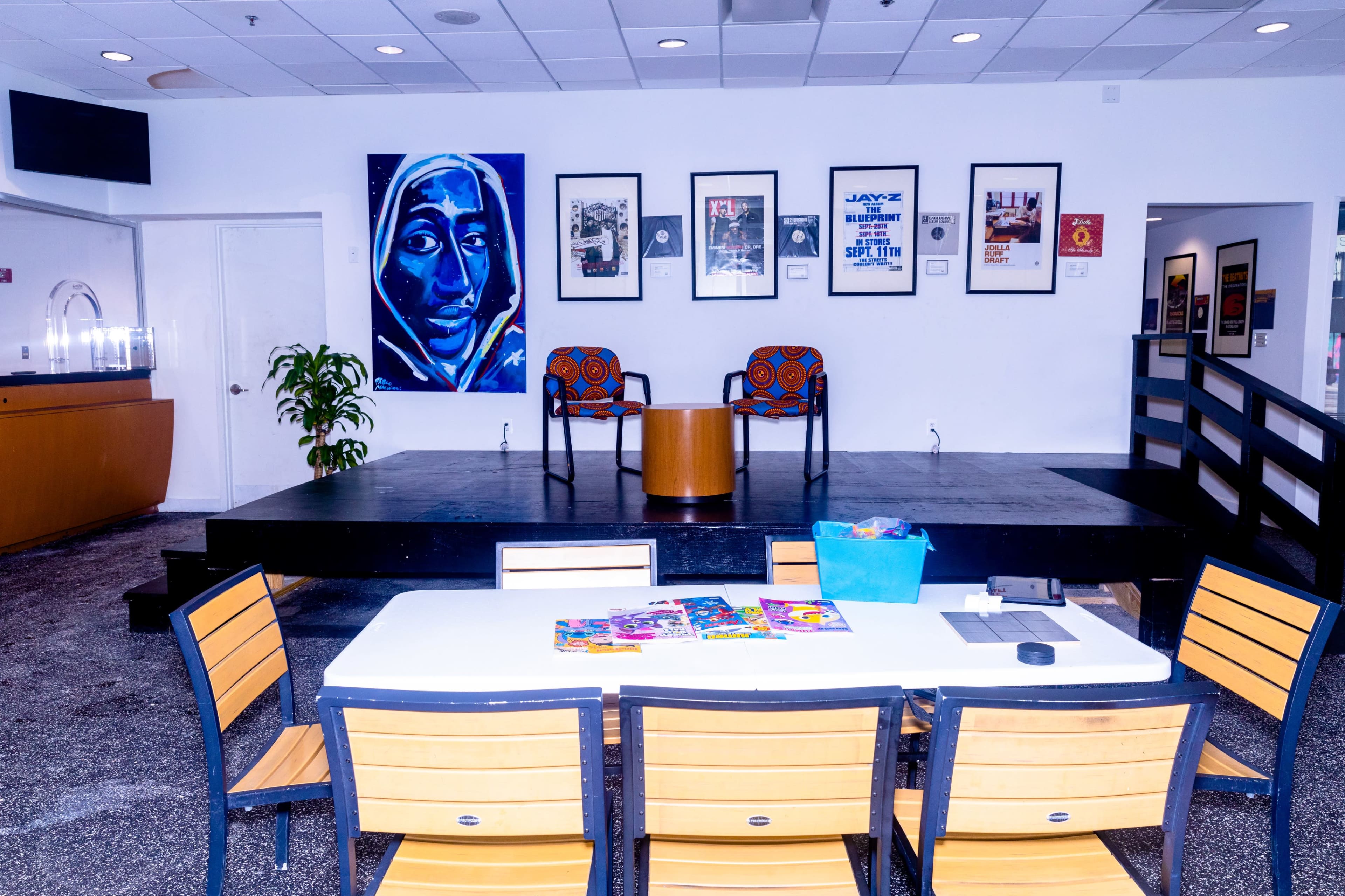 The image shows a bright community space with a table covered in art supplies, a small stage in the background, and framed artwork on the walls.