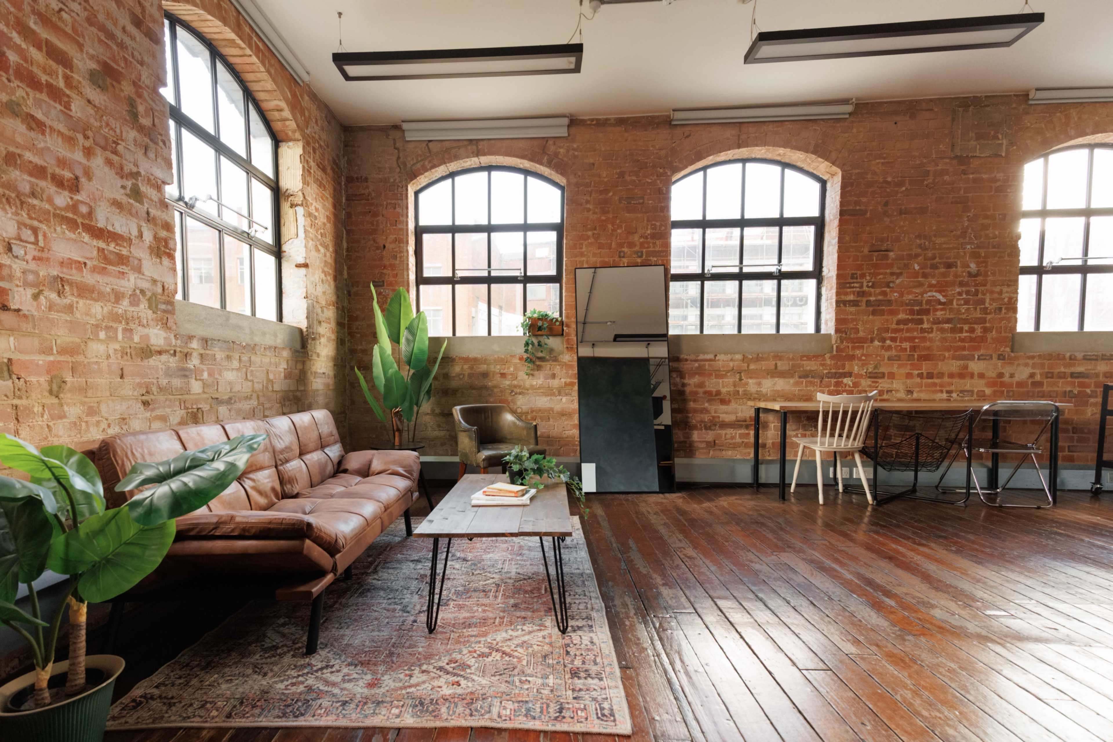 The image shows an industrial-style room with exposed brick walls, large windows, a brown leather sofa, a wooden coffee table, and a dining area with a mix of chairs.