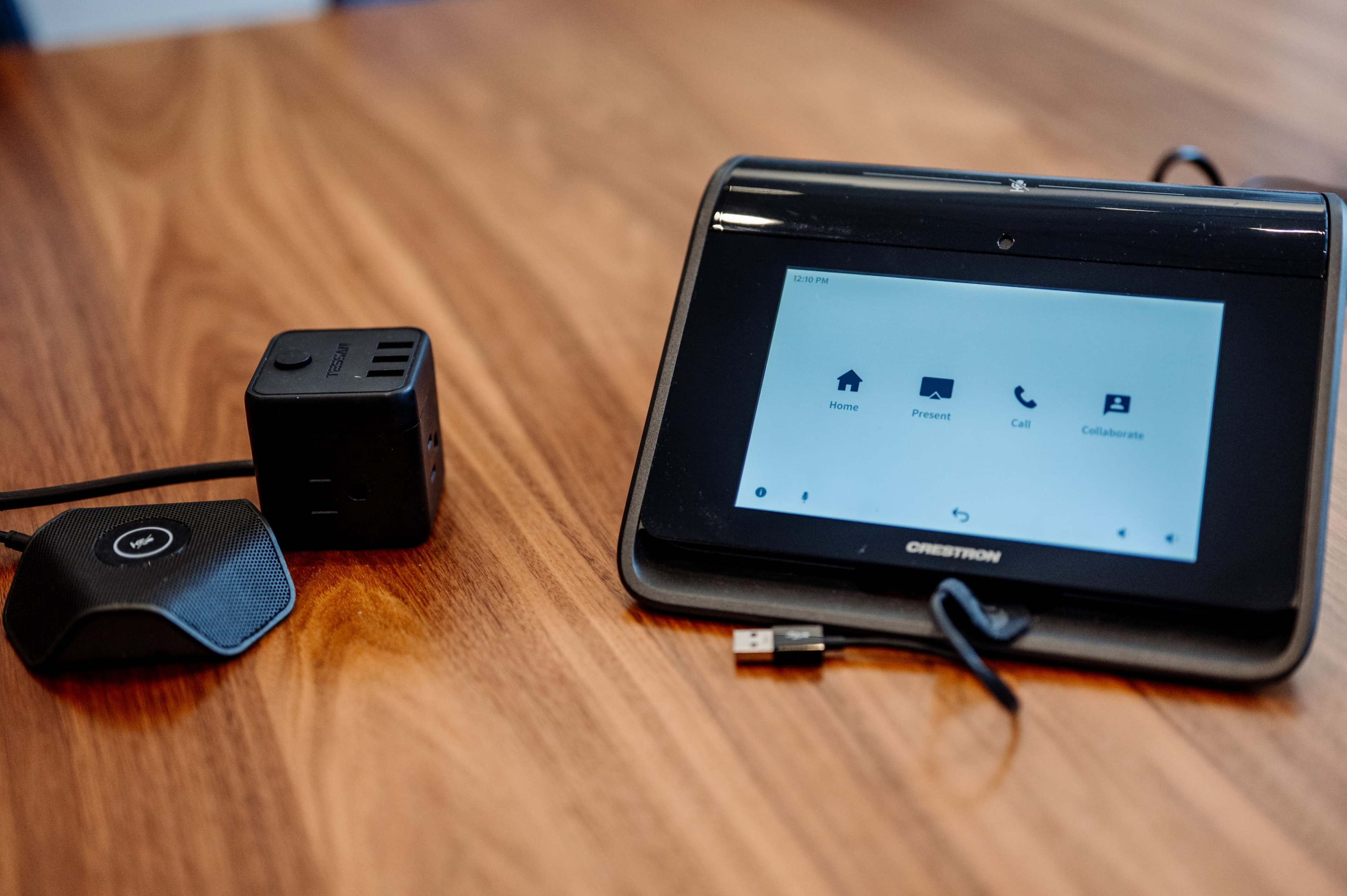 A Crestron touchscreen control panel sits on a wooden table next to a small black device with USB and power outlets.