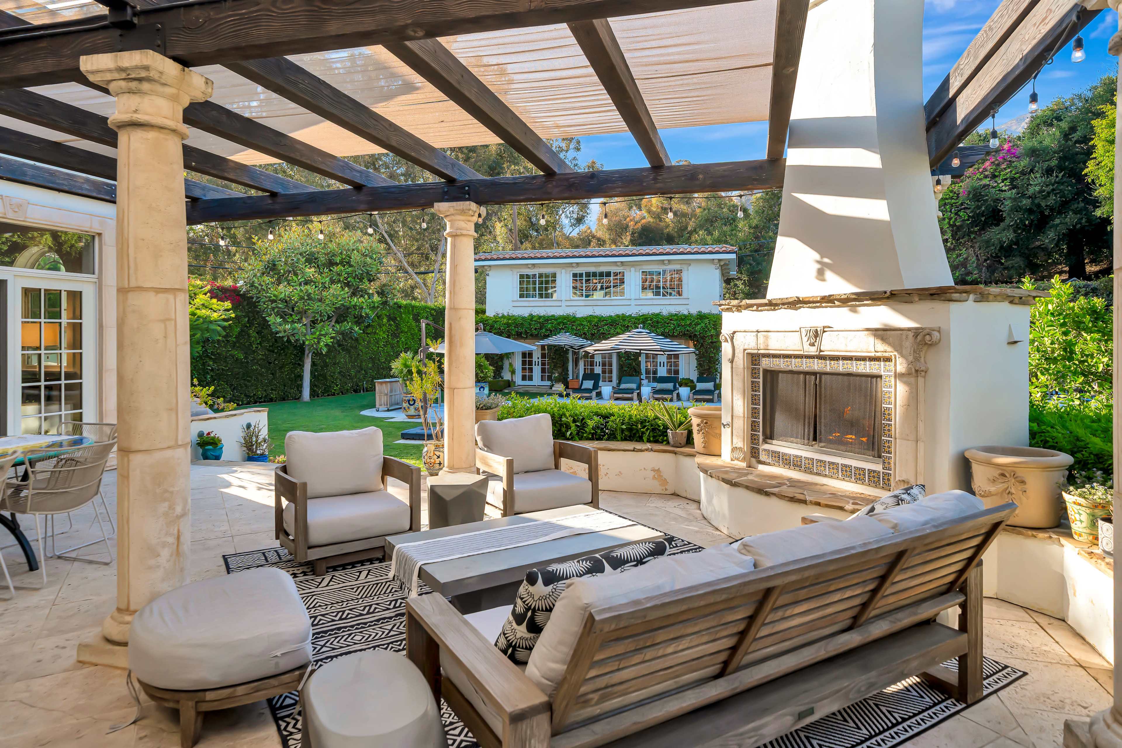 The image shows a luxurious outdoor seating area with a fireplace, overlooking a landscaped backyard featuring a pool and lounge chairs.