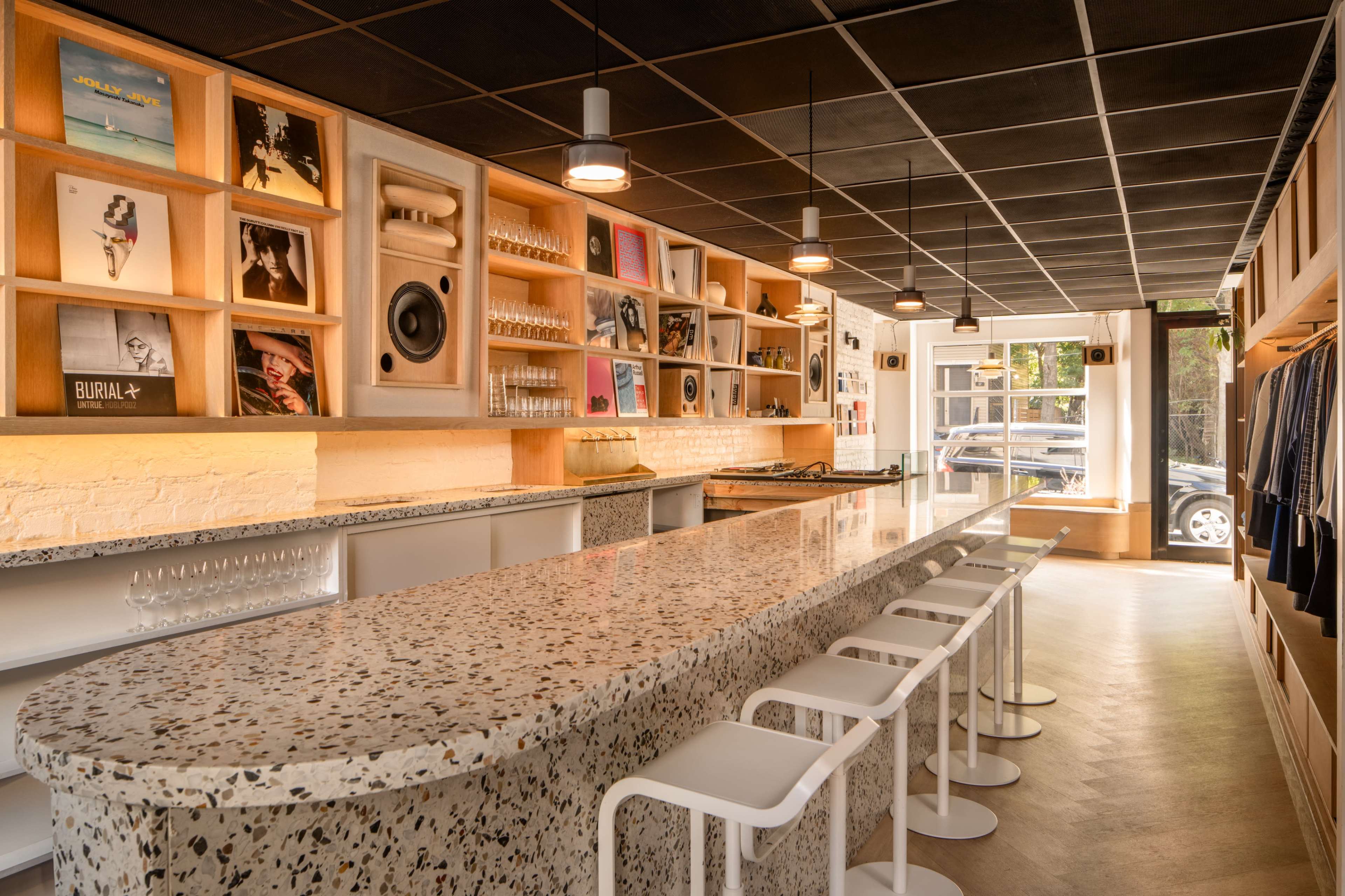 The image shows a modern, minimalist café with a long terrazzo bar, white stools, and wall displays featuring framed artwork and vinyl records.