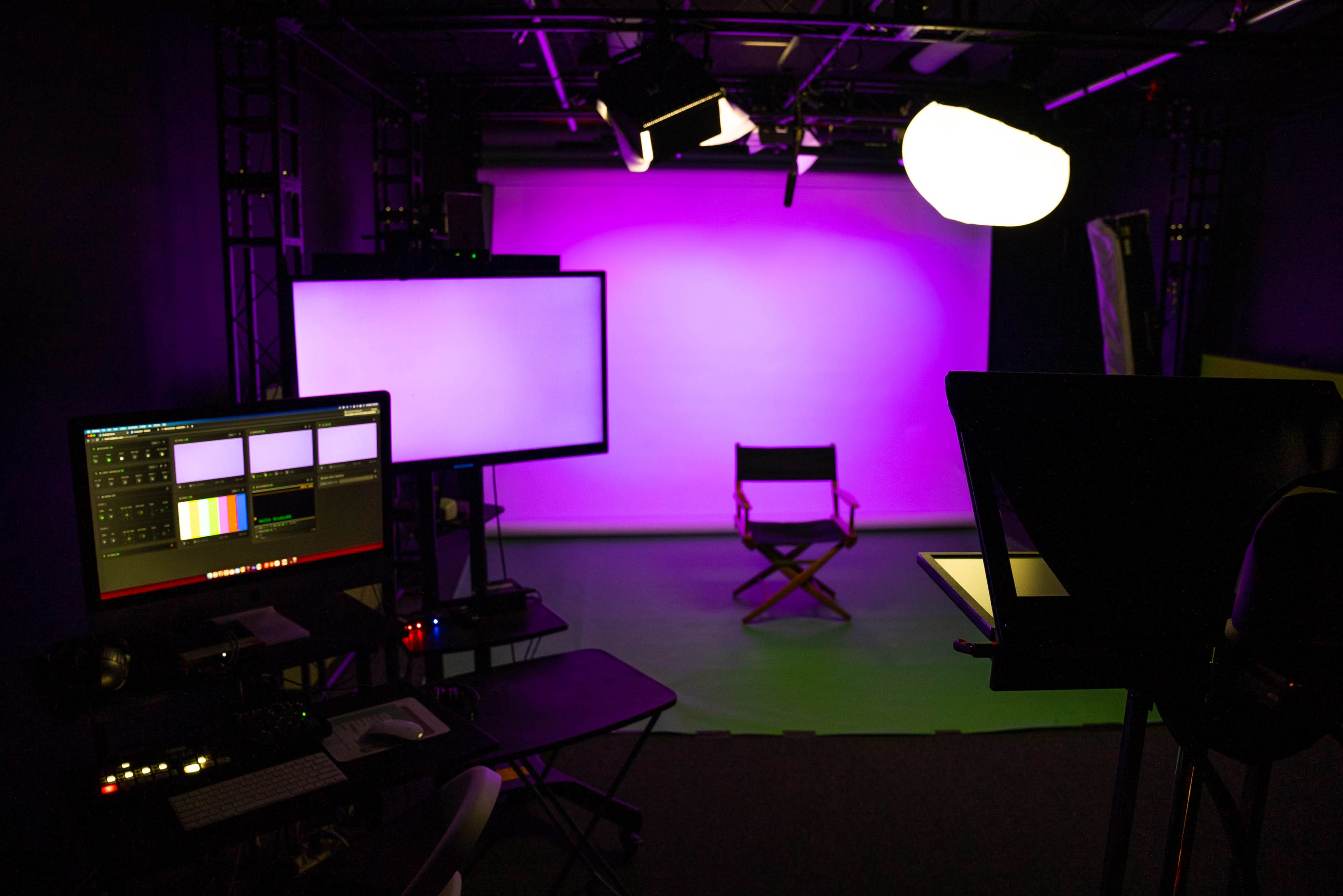 A video production studio is set up with a purple backdrop, a director's chair, and various screens and lighting equipment.
