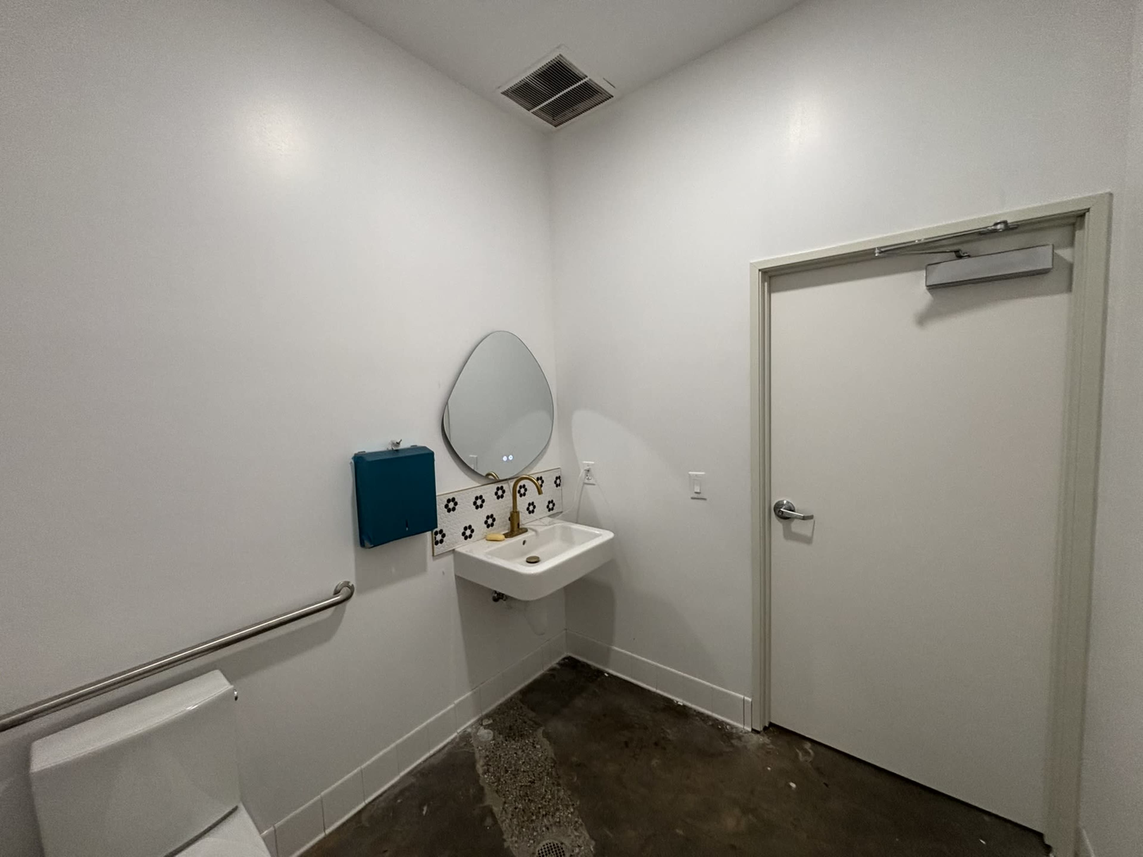 The image shows a small restroom with a wall-mounted sink, a mirror, a paper towel dispenser, and a white door.