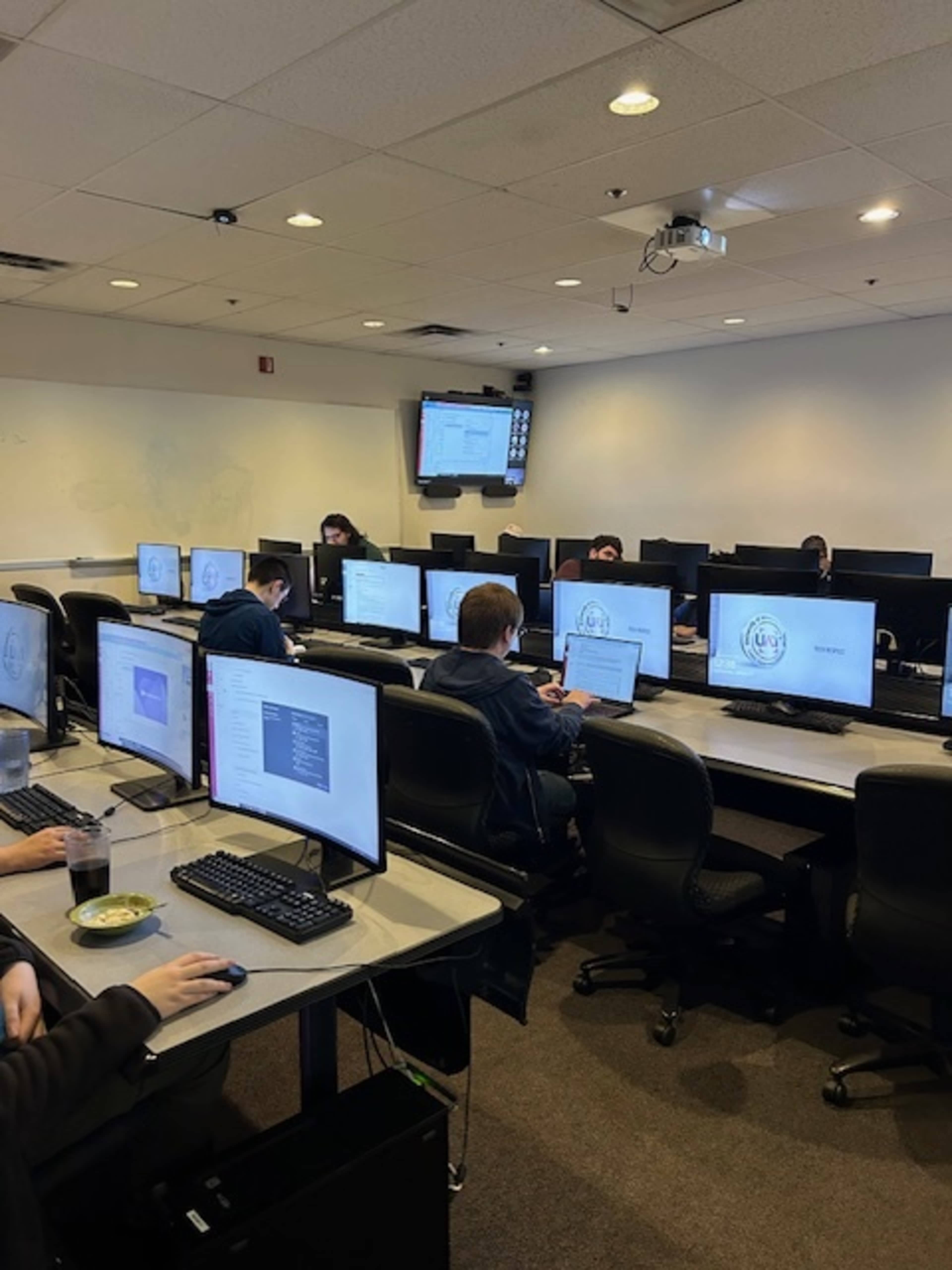 Several students work on computers in a classroom setting equipped with multiple desktop monitors.