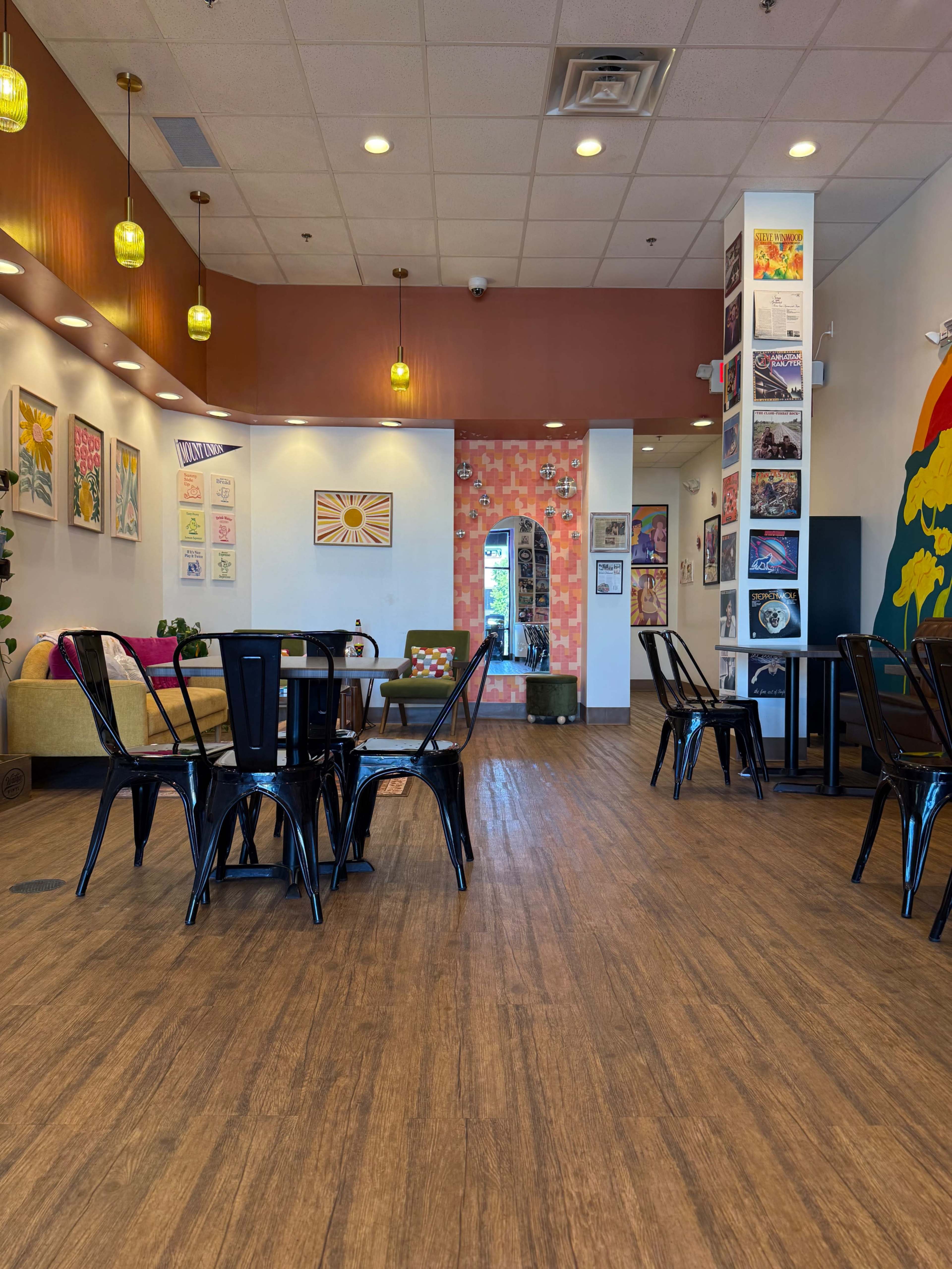 The image shows a brightly lit café with wooden flooring, metal chairs, and a mix of colorful artwork displayed on the walls.