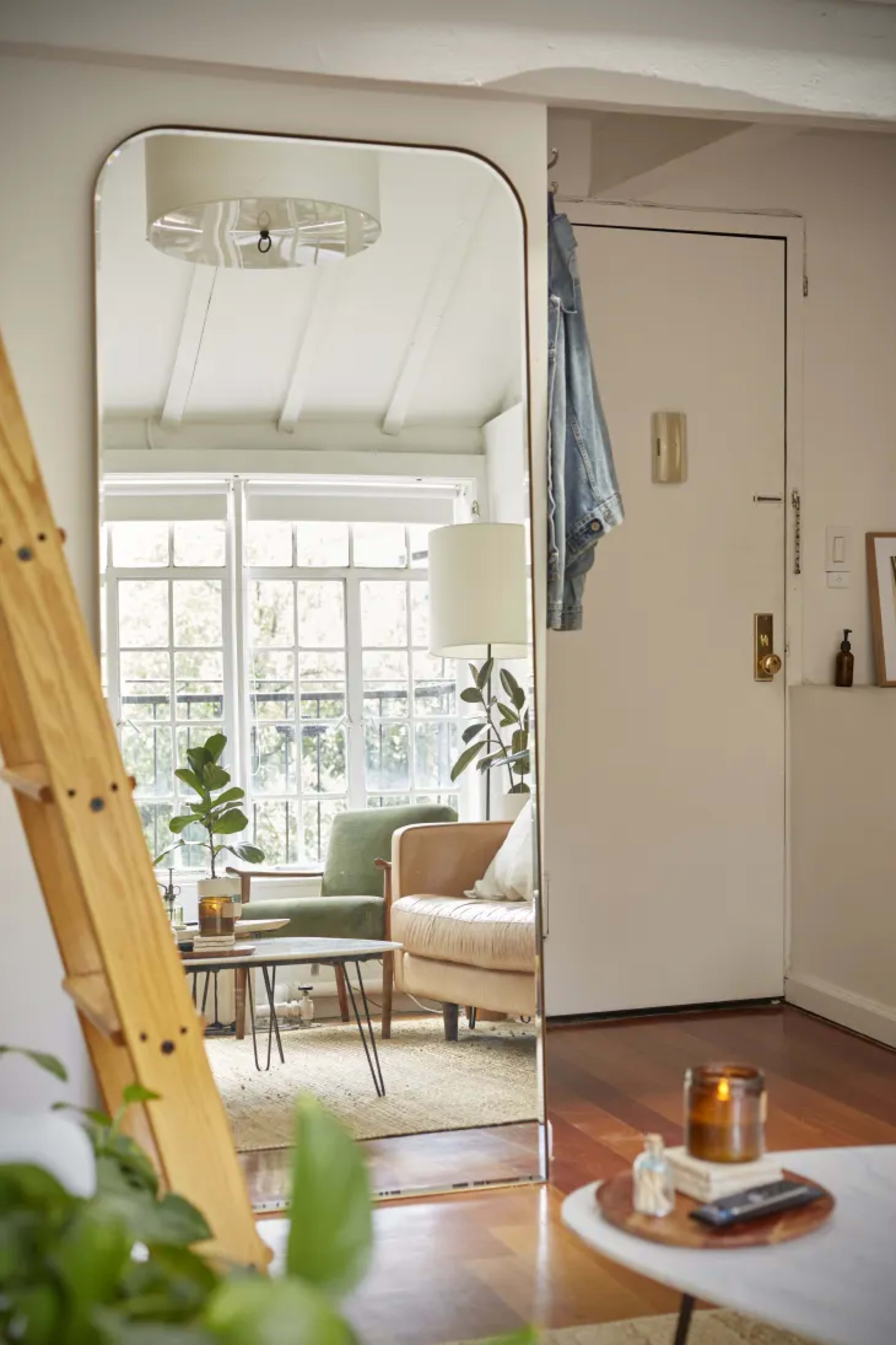 A tall mirror reflects a living space that includes a beige couch and several potted plants, with a door and window in the background.