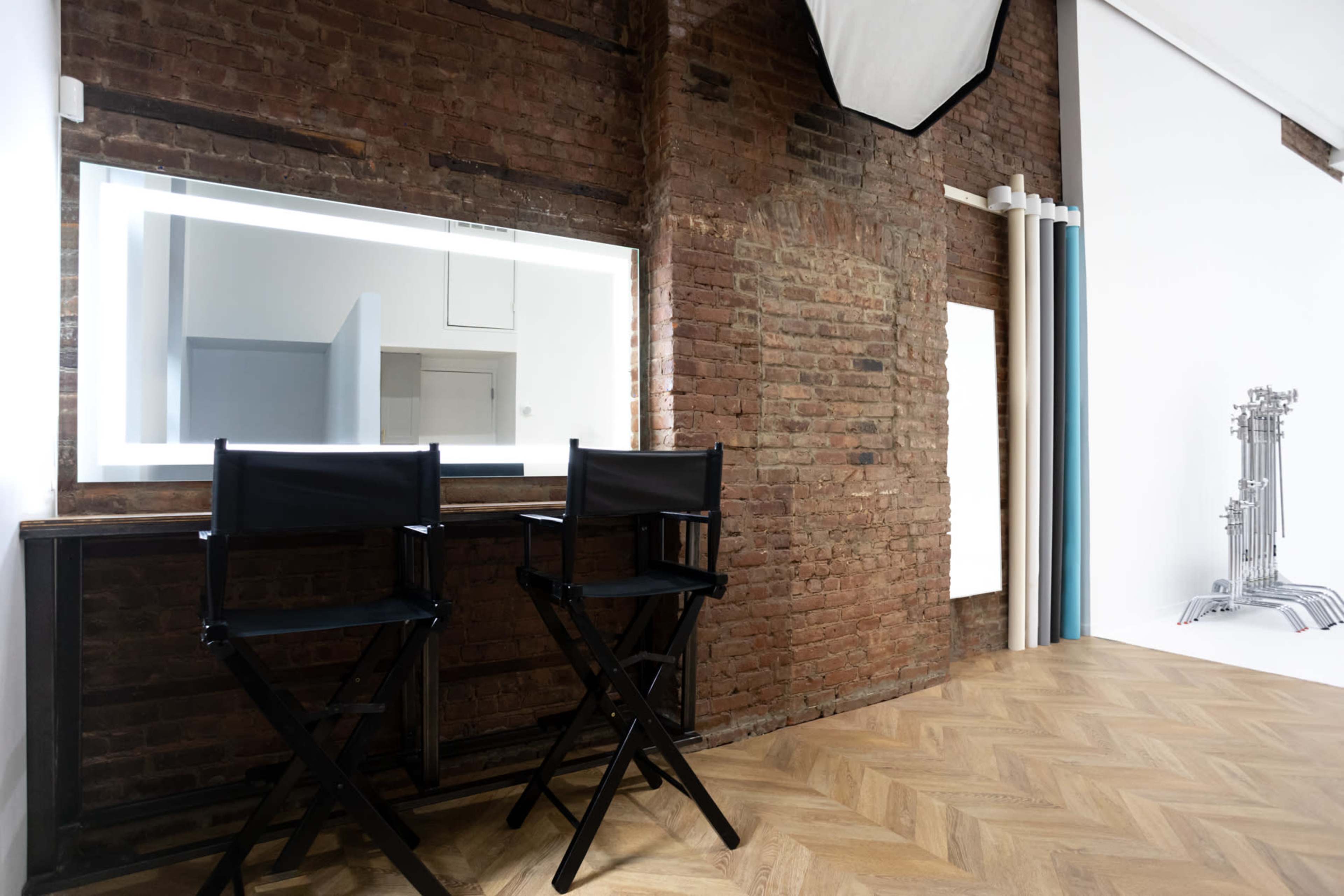 The image shows a minimalist interior featuring two black director's chairs at a wooden table next to a well-lit mirror mounted on a brick wall.