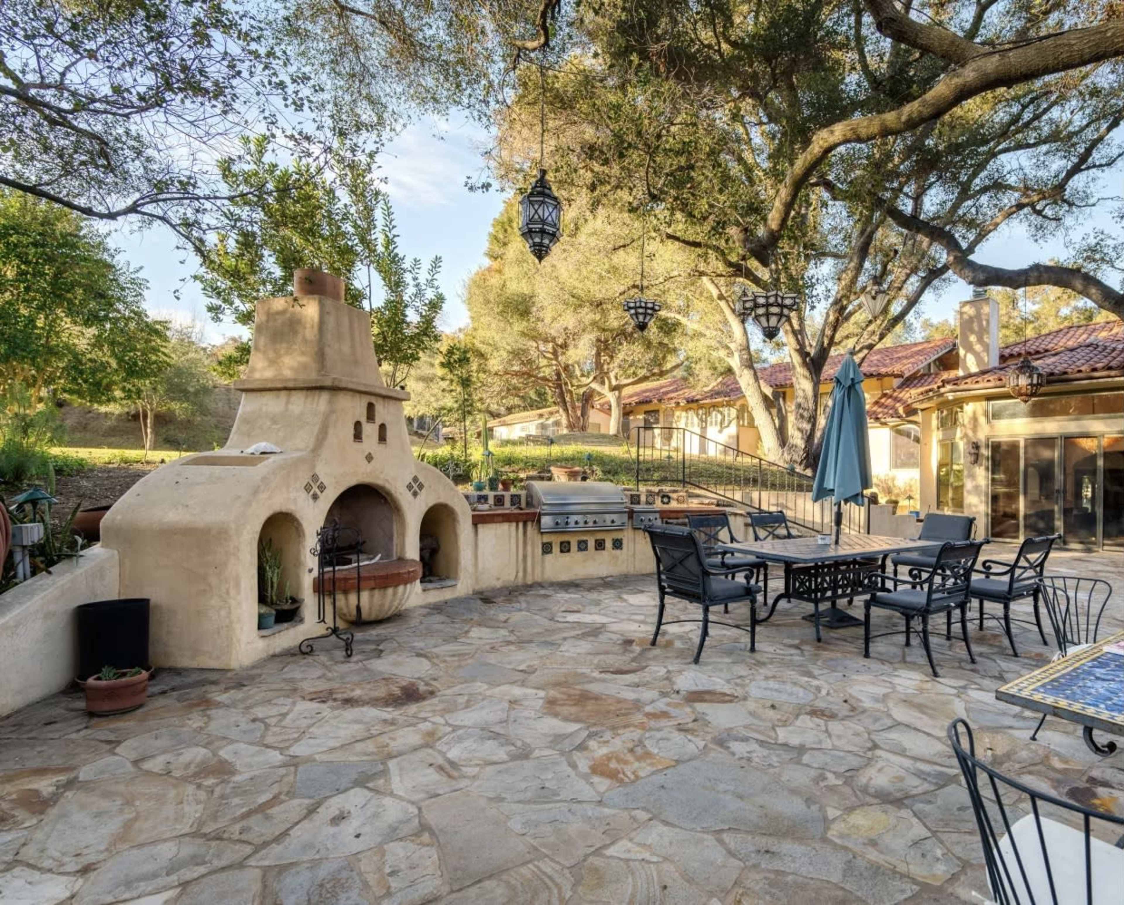 The image shows a spacious outdoor patio area featuring a large adobe-style fireplace, stone flooring, and black metal furniture surrounded by trees and decorative lanterns.