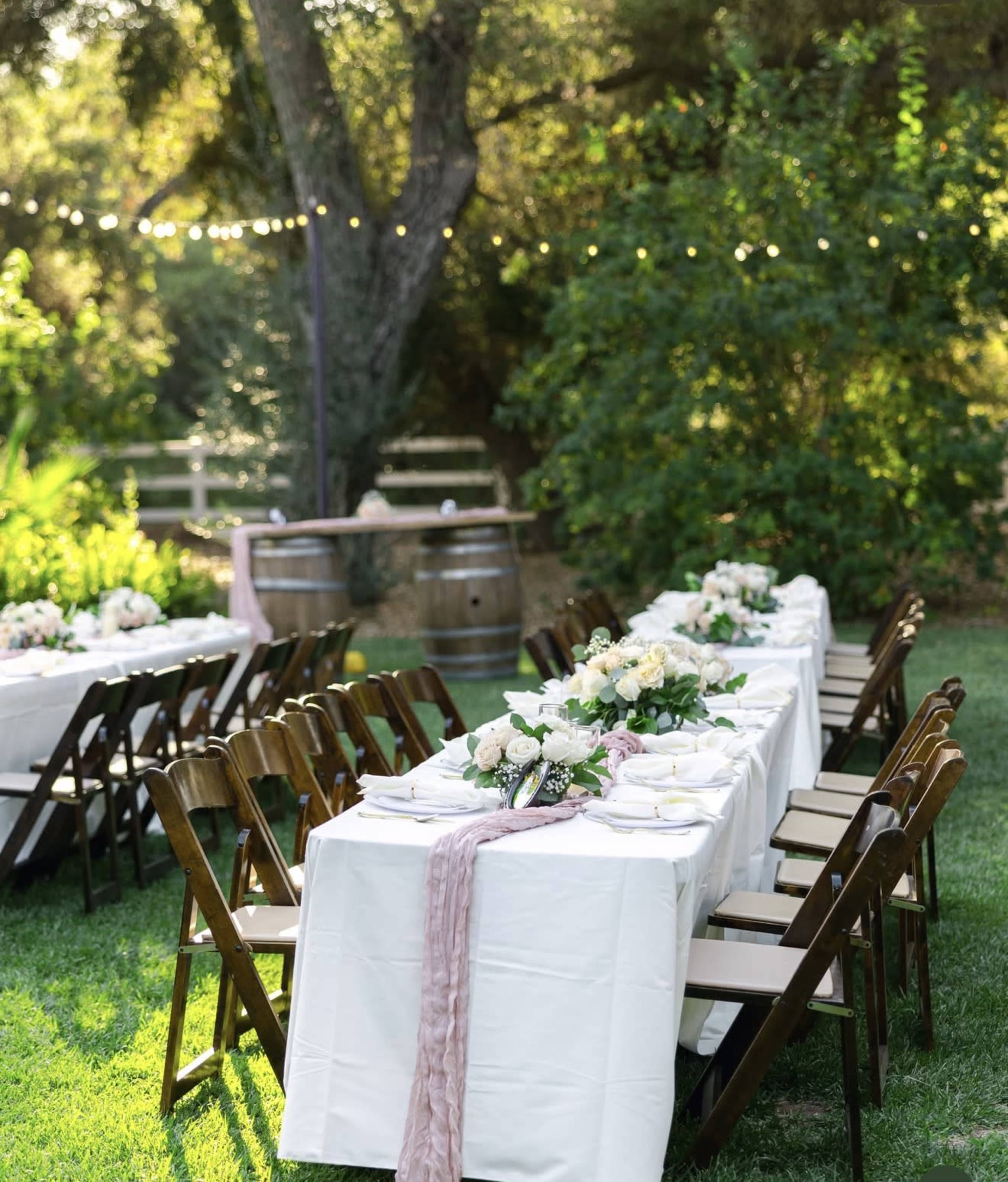 A long outdoor dining table is set with white tablecloths and floral centerpieces, surrounded by wooden chairs in a garden setting with string lights overhead.