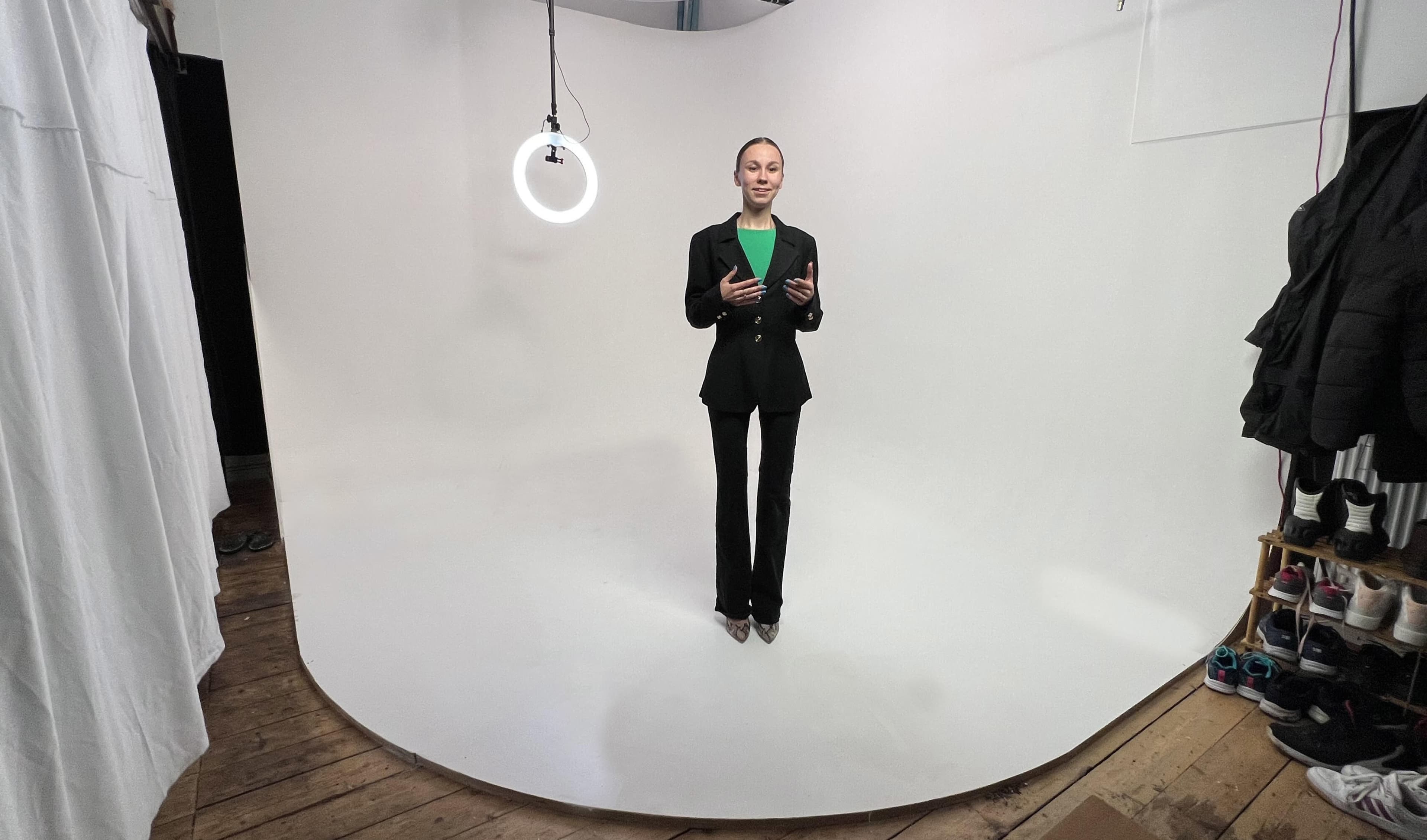 A woman in a black suit stands on a white backdrop, with a ring light suspended above her.