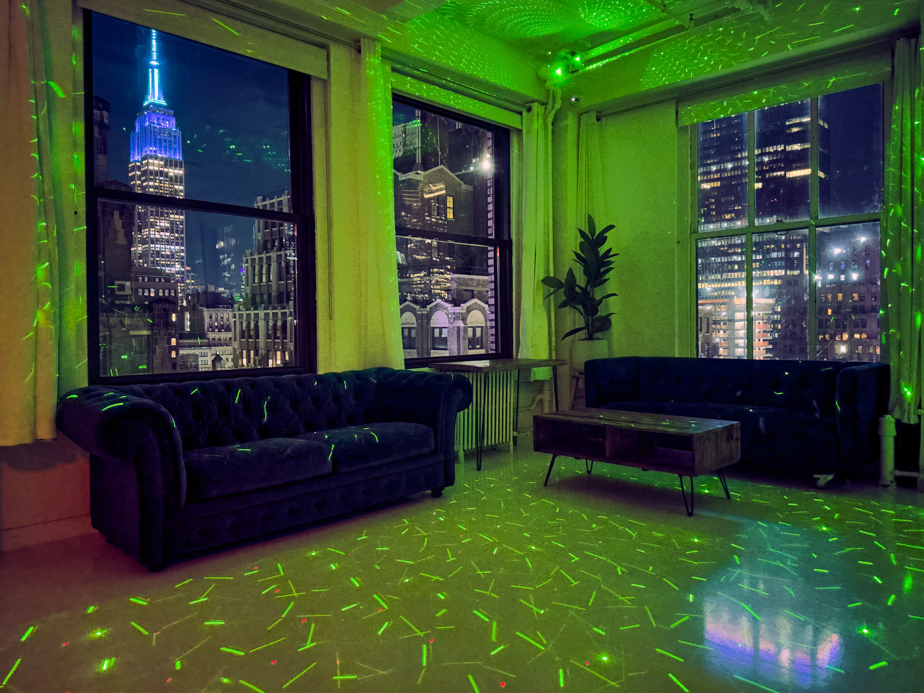 A spacious room with green laser lights projected on the floor and a view of the Empire State Building through large windows.