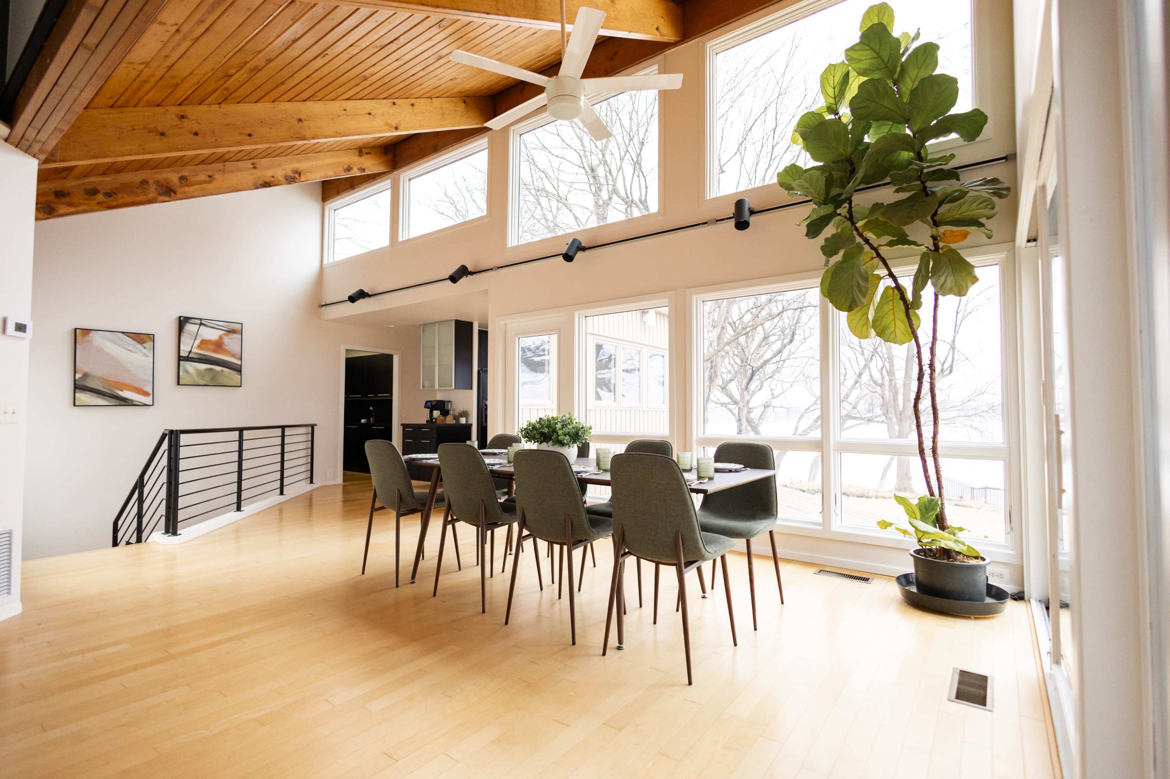 The image shows a modern dining area with a large table set for six, surrounded by windows that provide a view of trees outside.