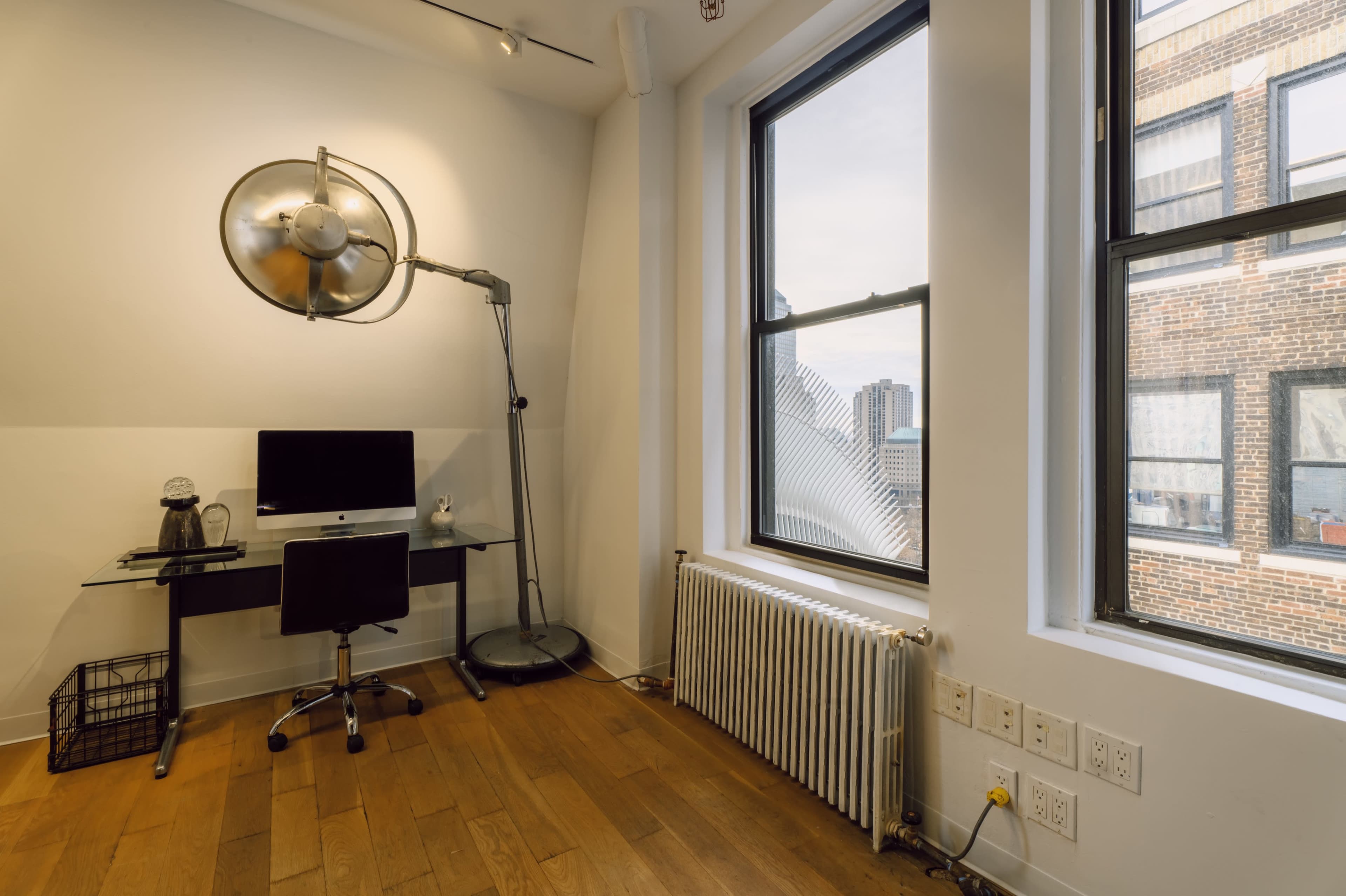 The image shows a modern workspace featuring a glass desk with a computer, a stylish industrial lamp, and a radiator beneath large windows revealing a cityscape.