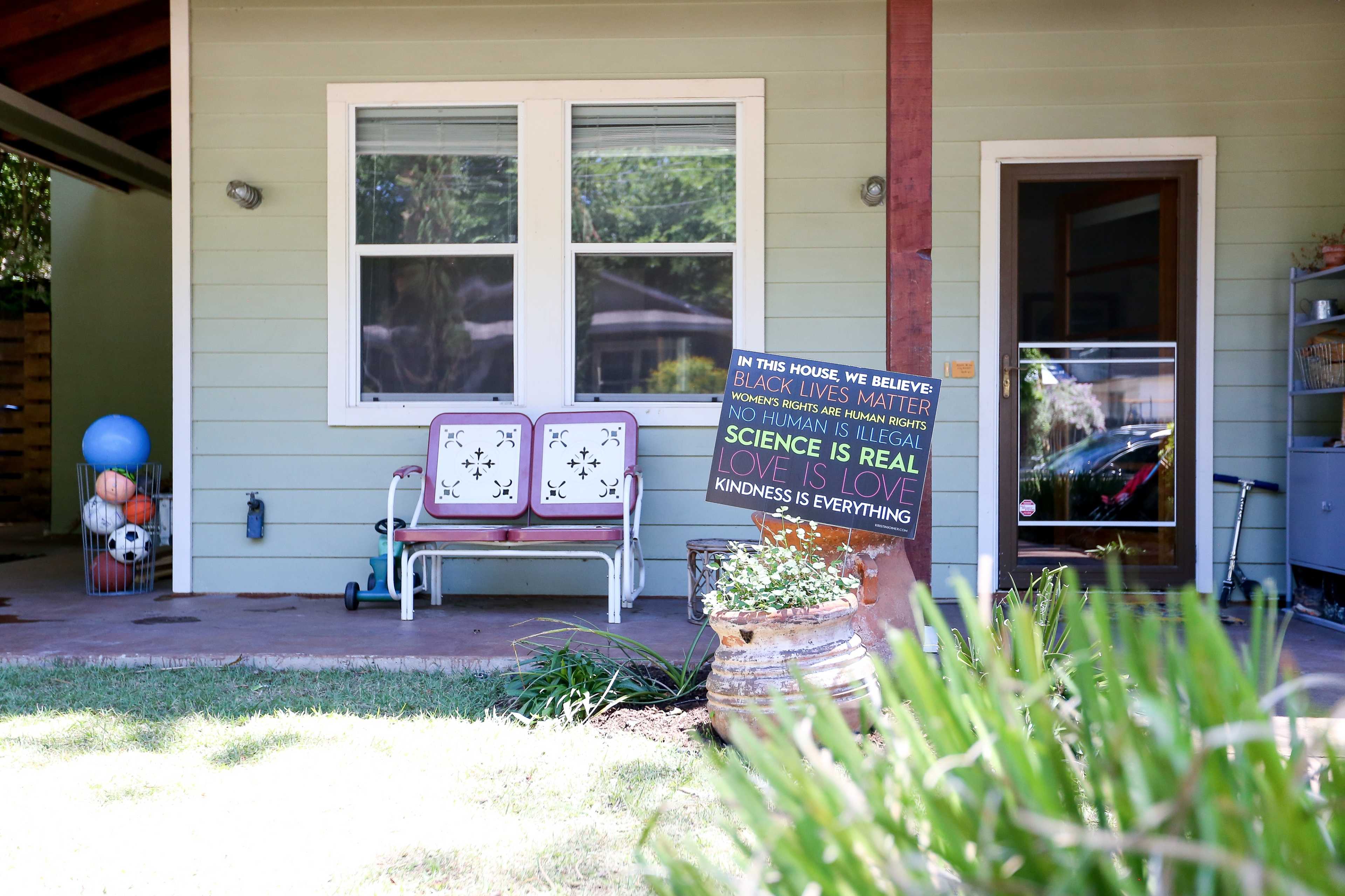 The image shows a house with a porch displaying a sign that includes several social justice messages and two chairs beside a small garden area.