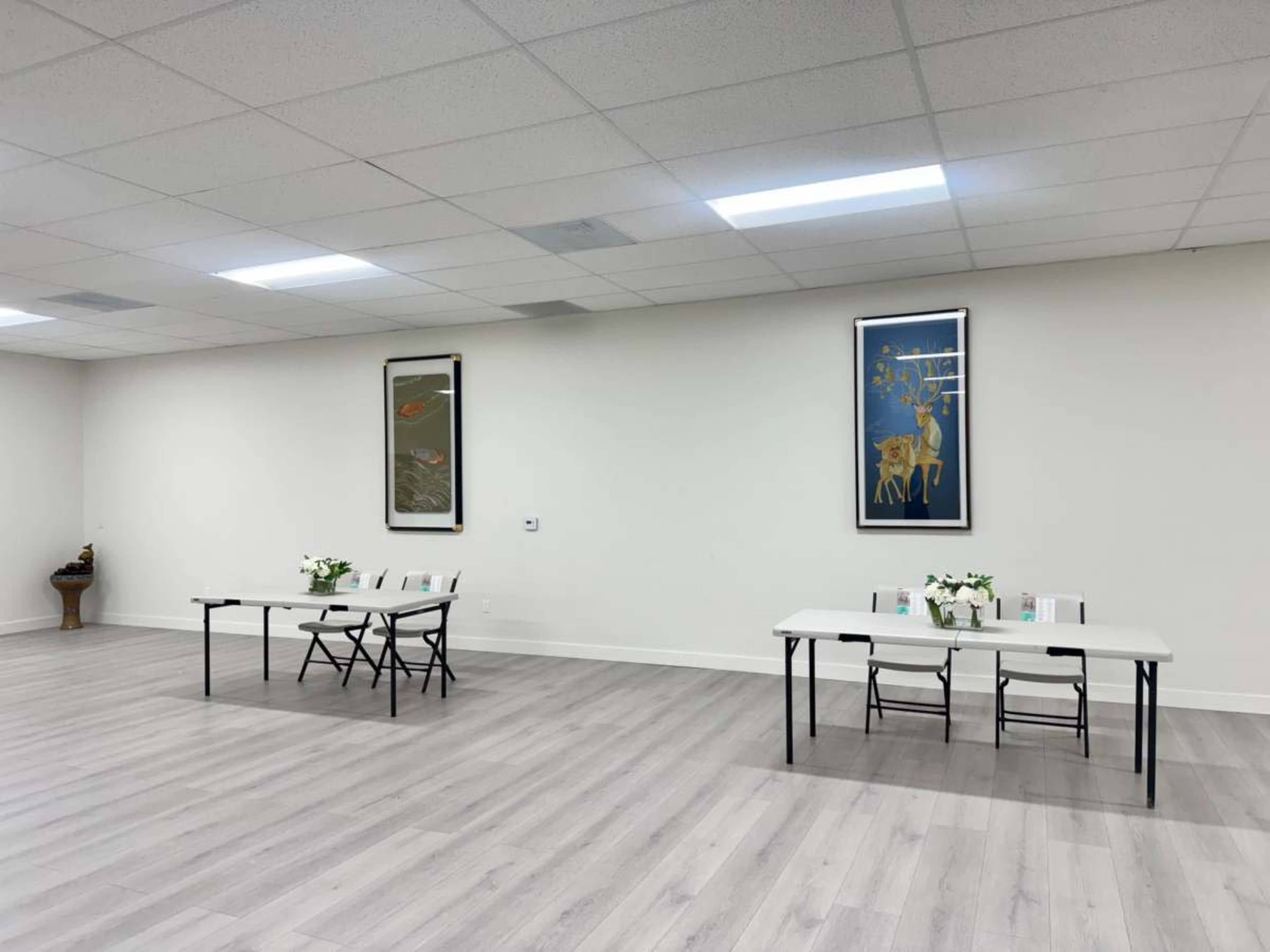The image shows a spacious, minimally decorated room with two tables set up, each adorned with flowers and books.
