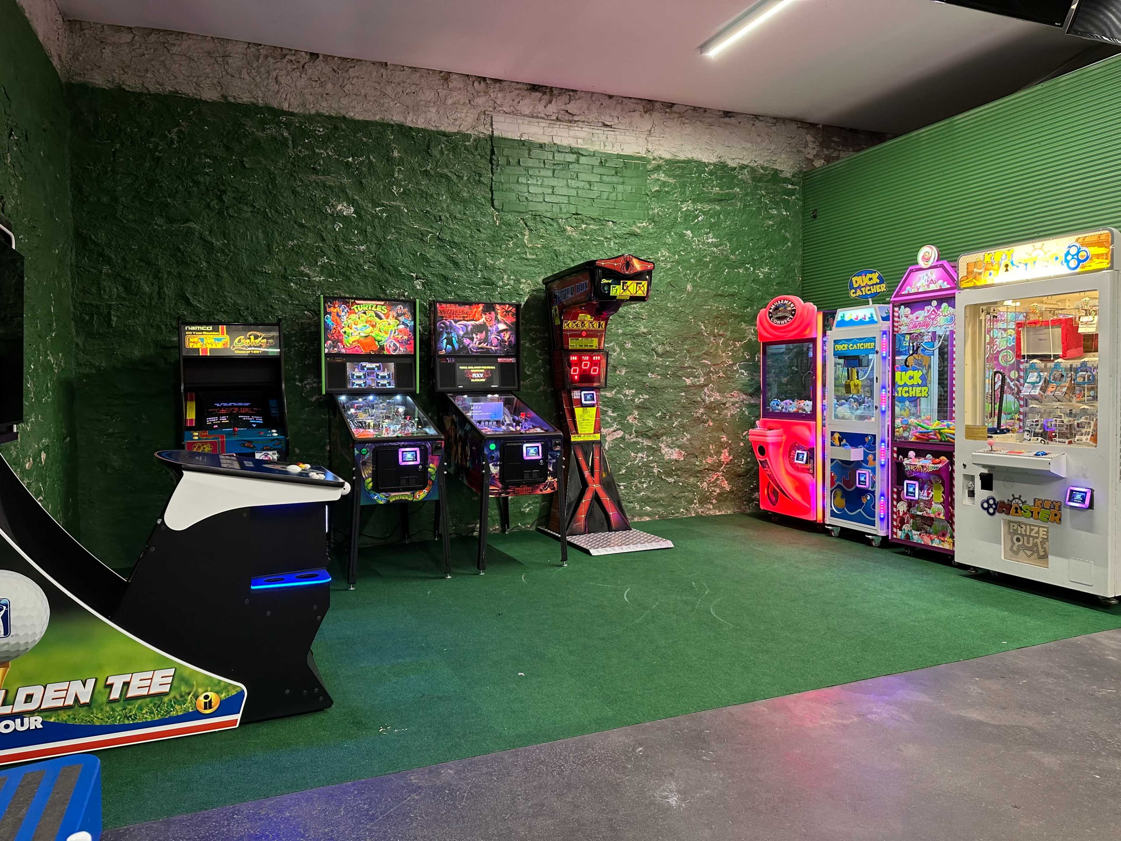 A corner of an arcade with various classic and modern gaming machines arranged against a green brick wall.