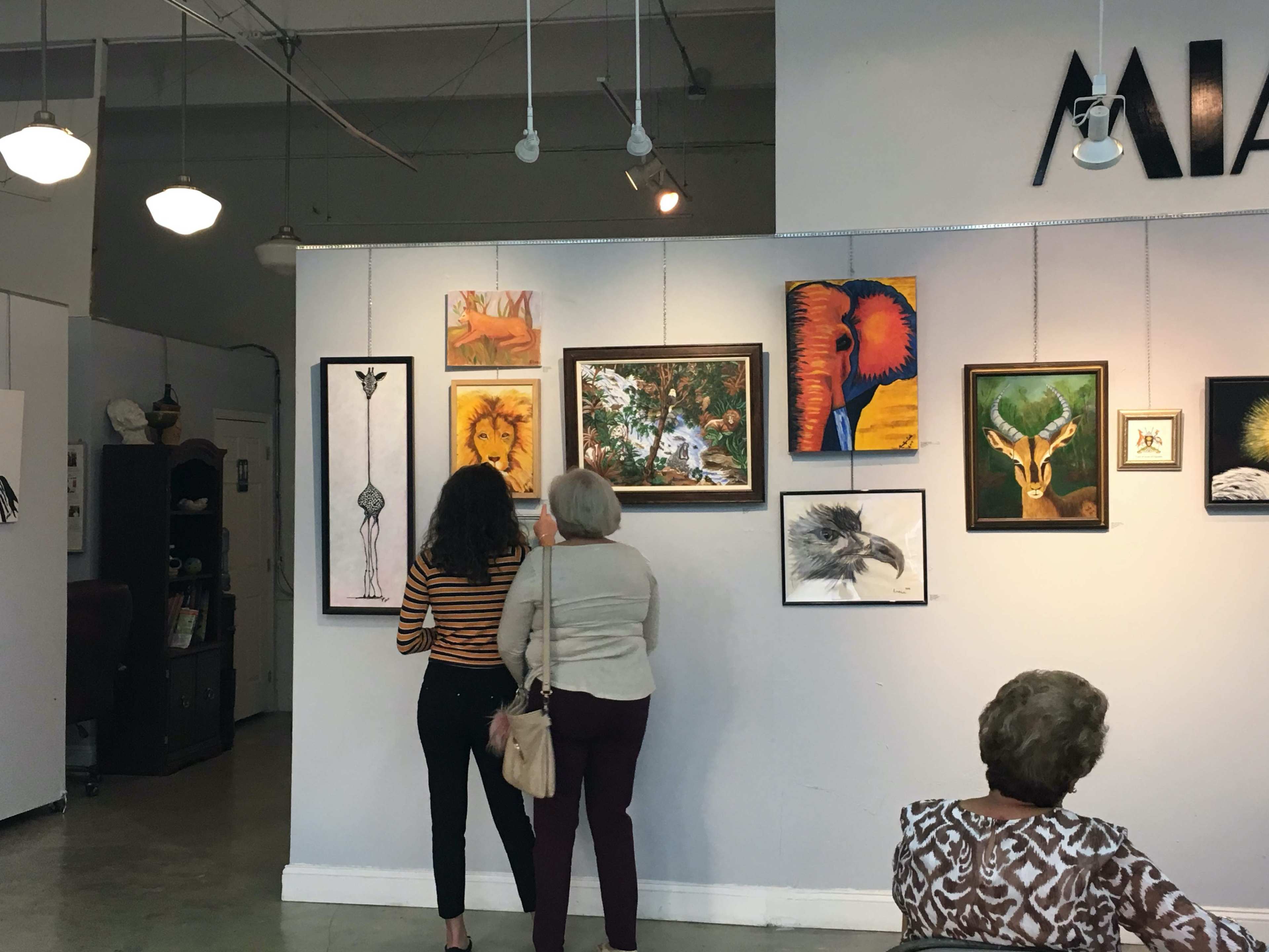Two women observe a collection of framed artwork displayed on a gallery wall, while another woman sits in the foreground.
