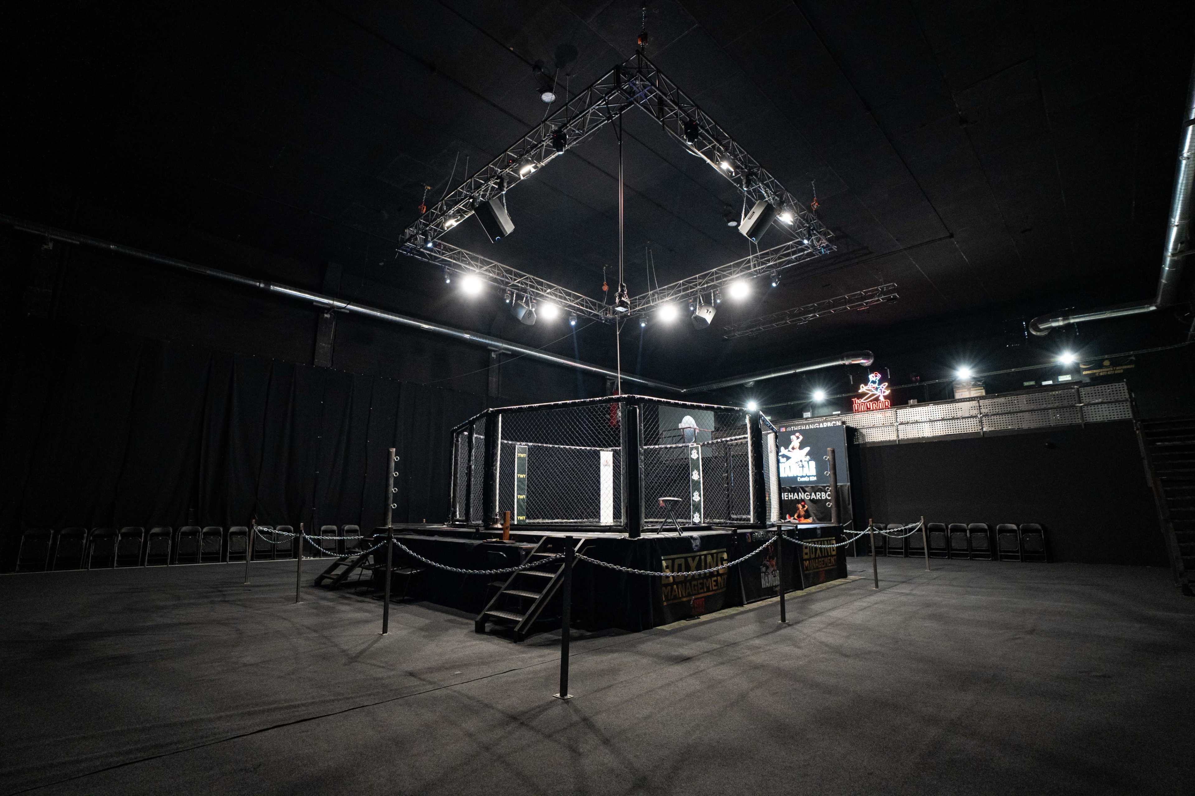 The image shows an empty indoor arena featuring a central octagonal cage surrounded by seating and illuminated by overhead lights.
