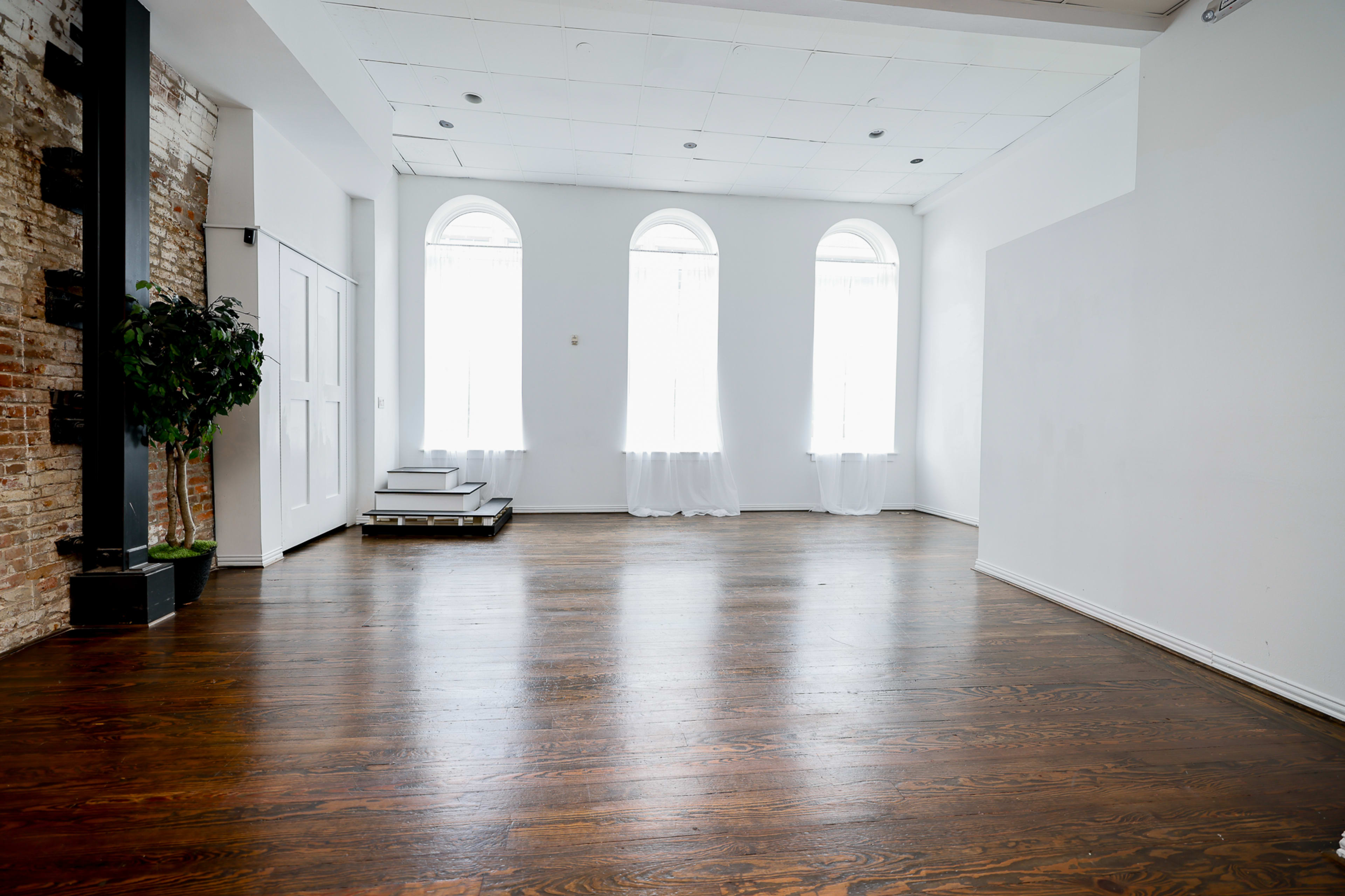 The image shows a spacious, well-lit interior with large arched windows, wooden floors, and minimal furnishings.