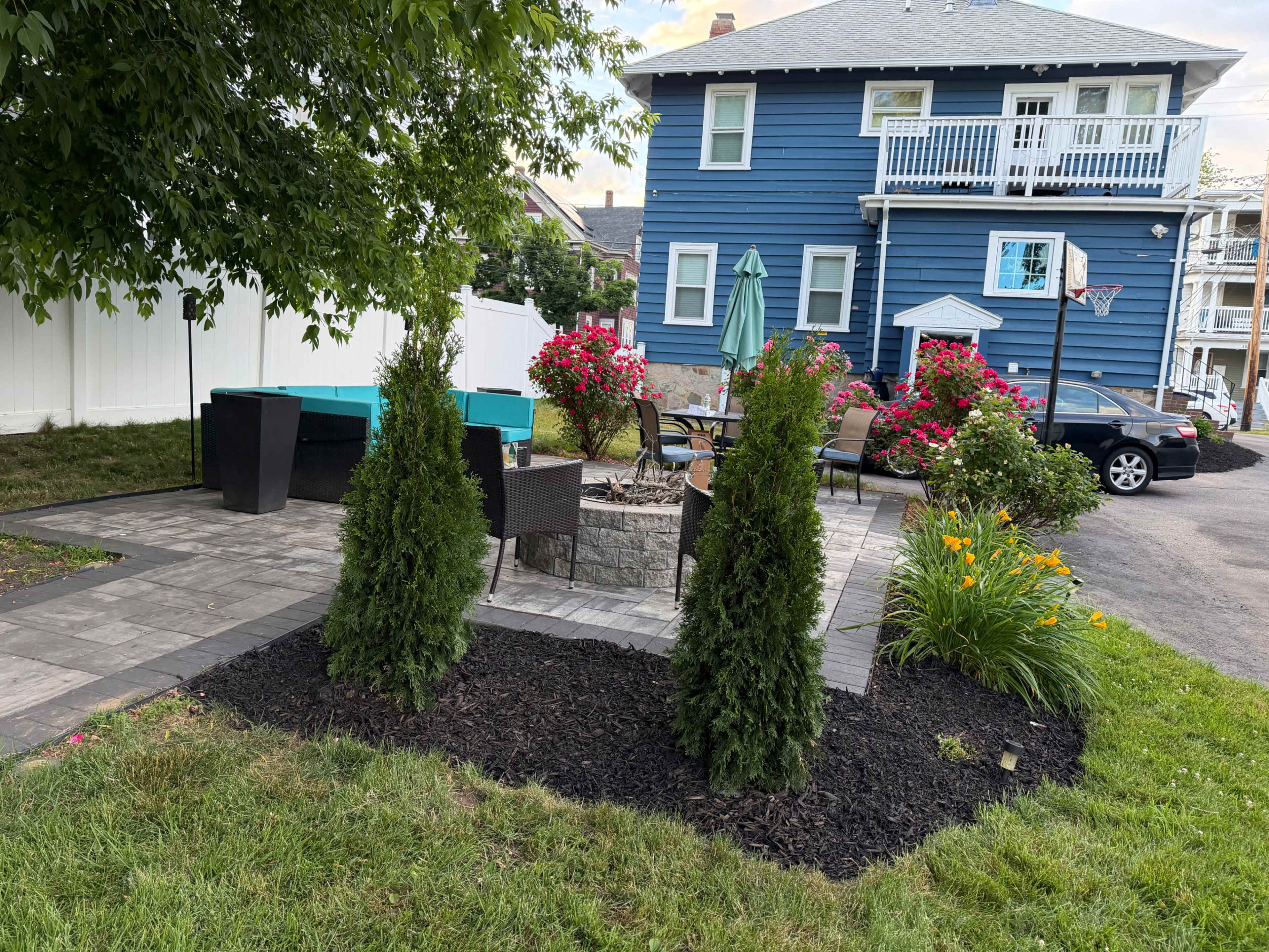 The image shows a landscaped outdoor patio area with seating, a fire pit, and colorful flower beds, adjacent to a blue house.