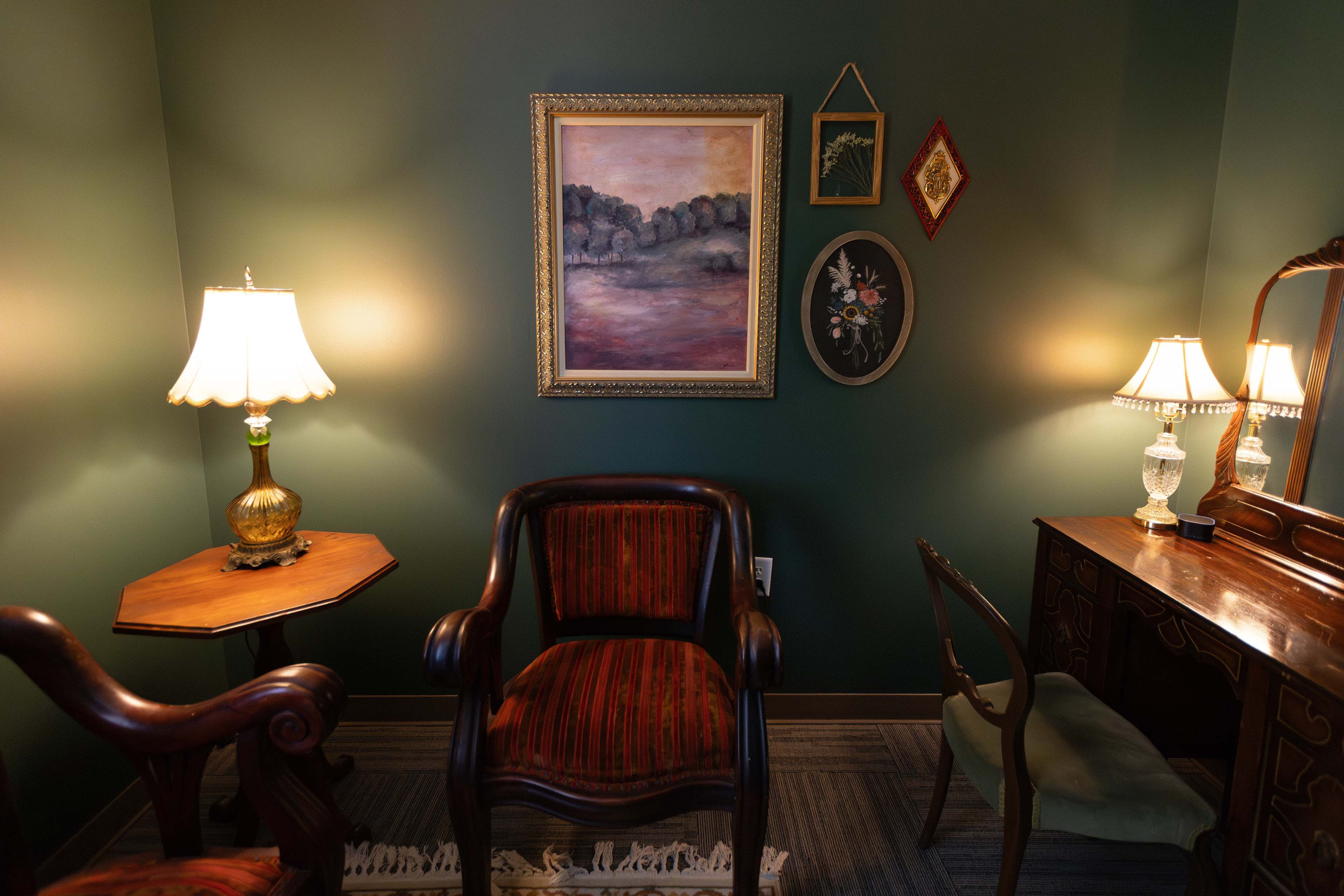 A warmly lit room featuring two lamps, a painting on the wall, and vintage-style furniture, including a wooden chair and a dresser.