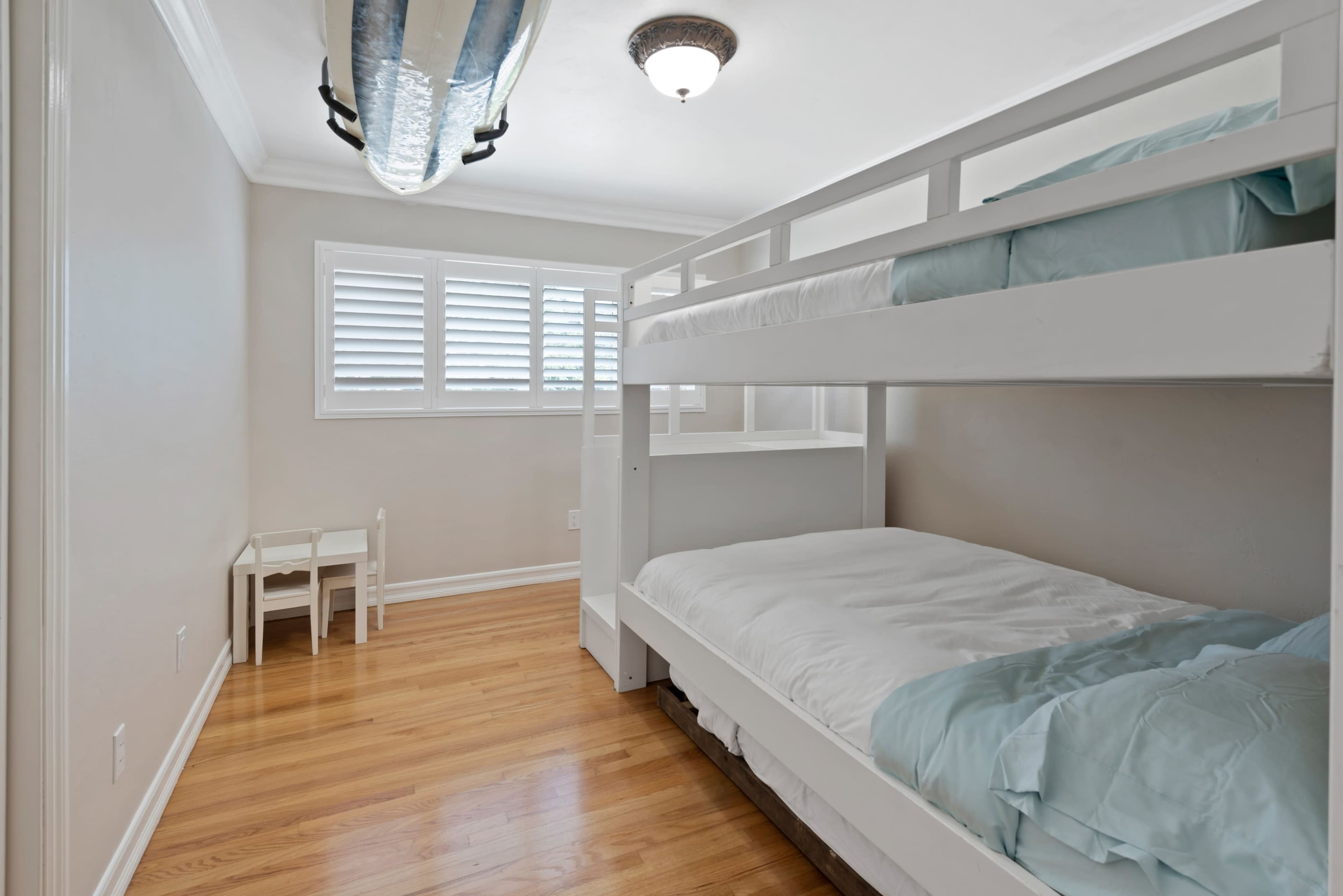 The image shows a well-lit bedroom featuring a bunk bed with light blue bedding and a small table and chairs against one wall.