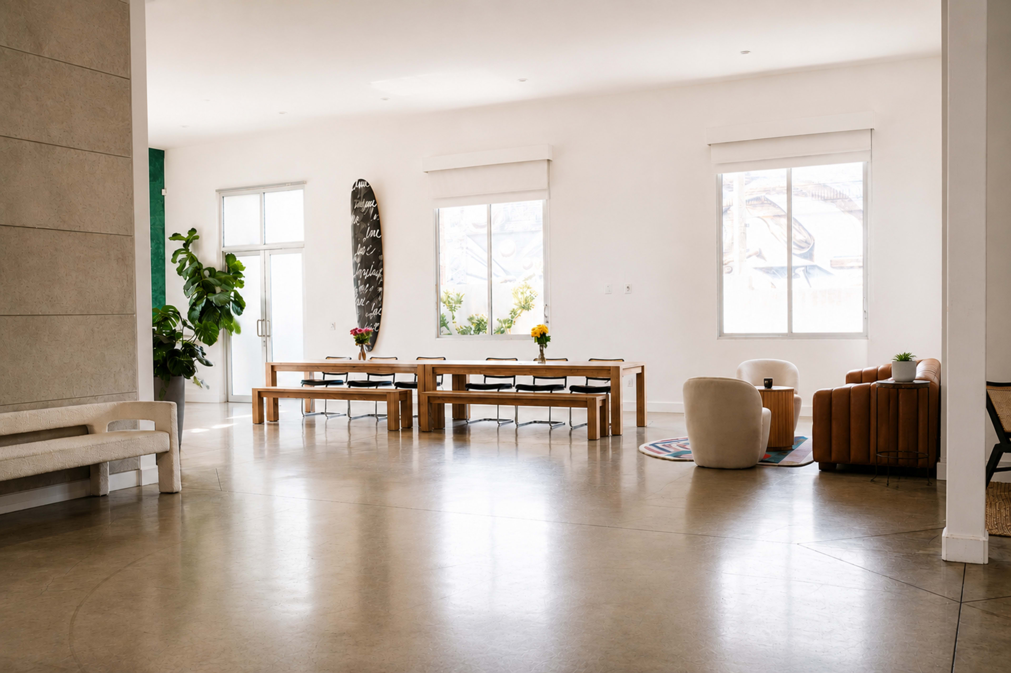 The image shows a spacious, well-lit modern interior featuring a large dining table with chairs, a comfortable seating area, a surfboard on the wall, and decorative plants.