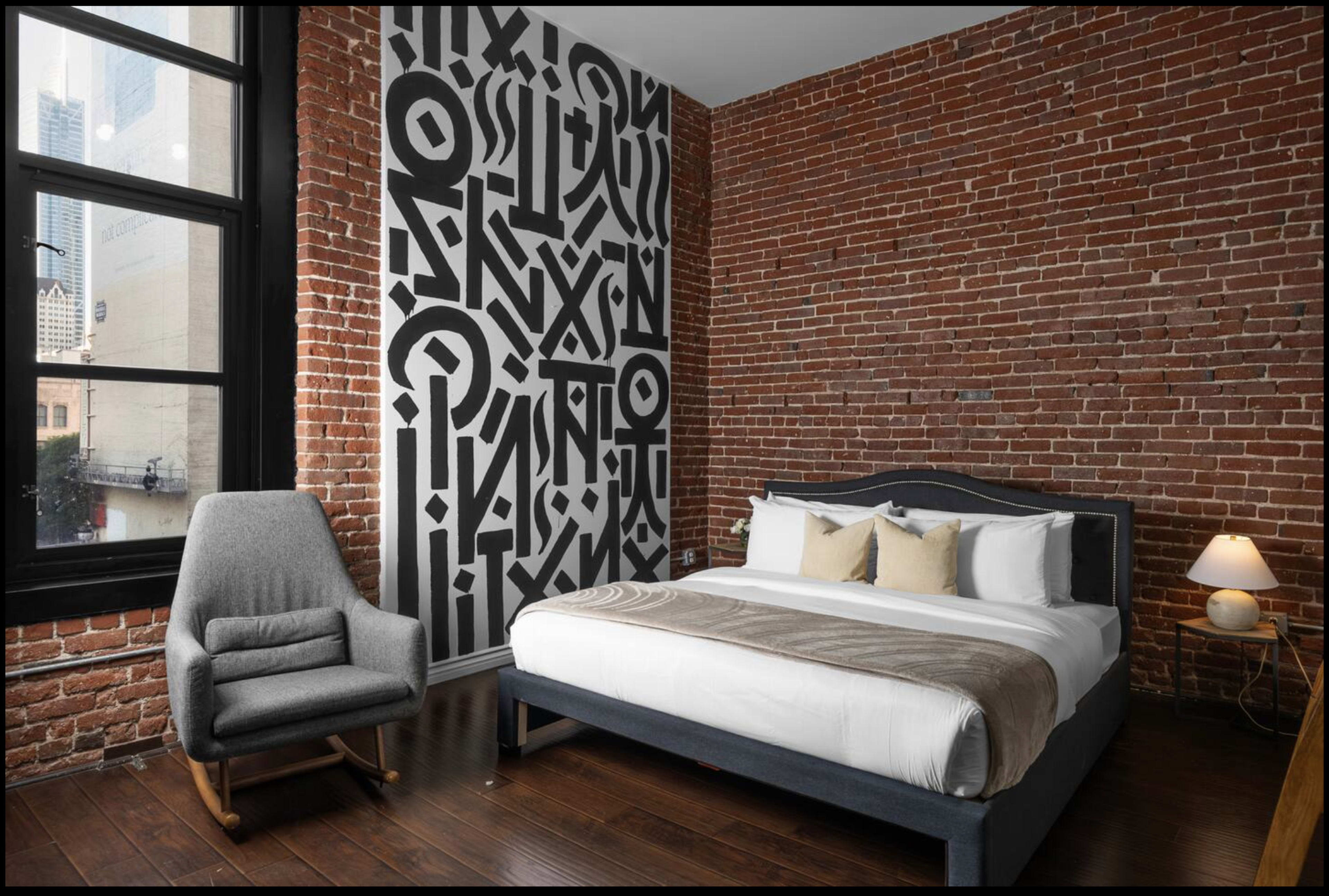 The image shows a modern bedroom featuring a double bed, exposed brick walls, a patterned accent wall, a gray armchair, a wooden nightstand, and a lamp.